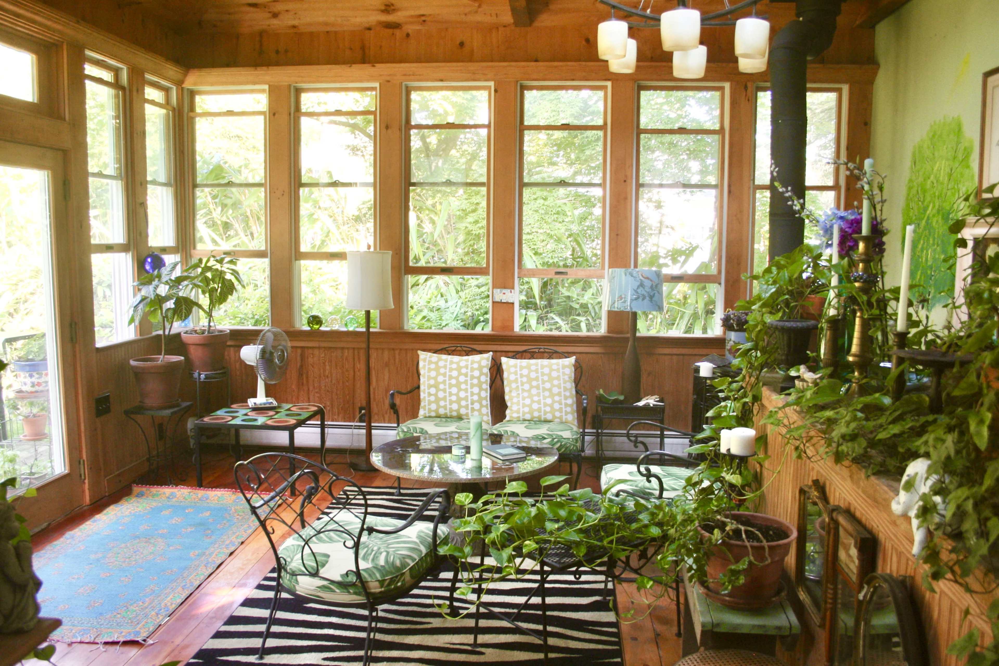 A sunlit room features a glass table surrounded by metal chairs, plants, and large windows overlooking a garden.