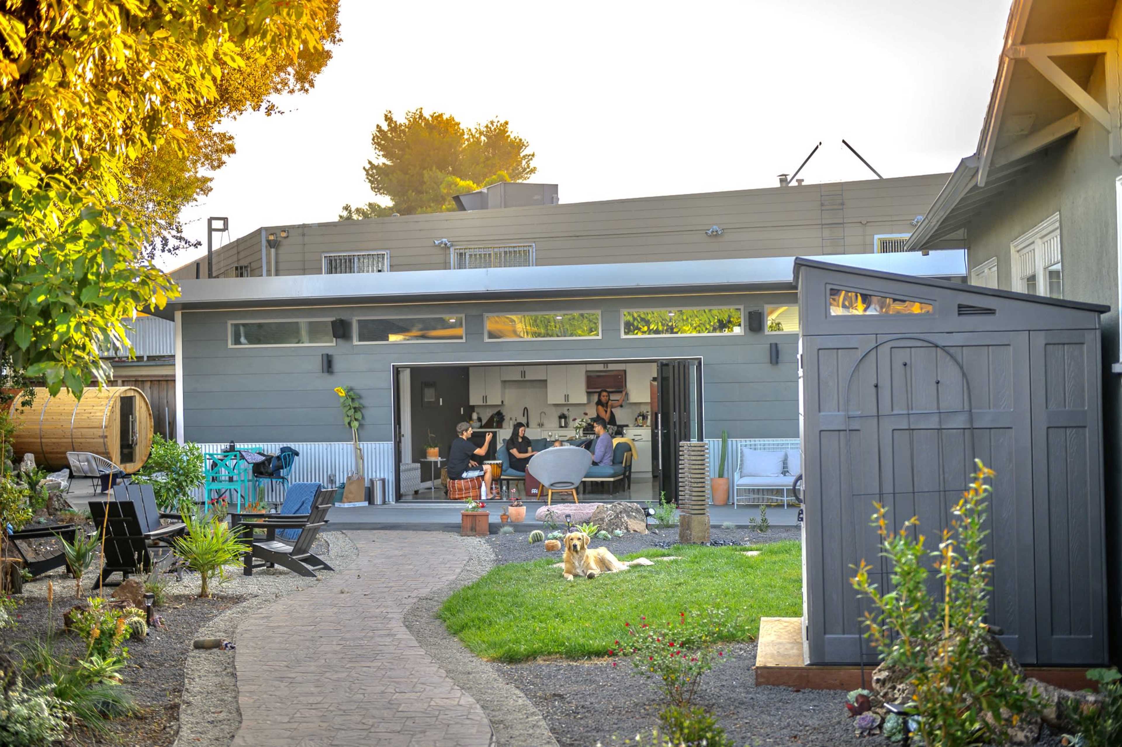 A modern backyard scene features a home with large sliding glass doors opening to a living area, surrounded by greenery and garden decor.
