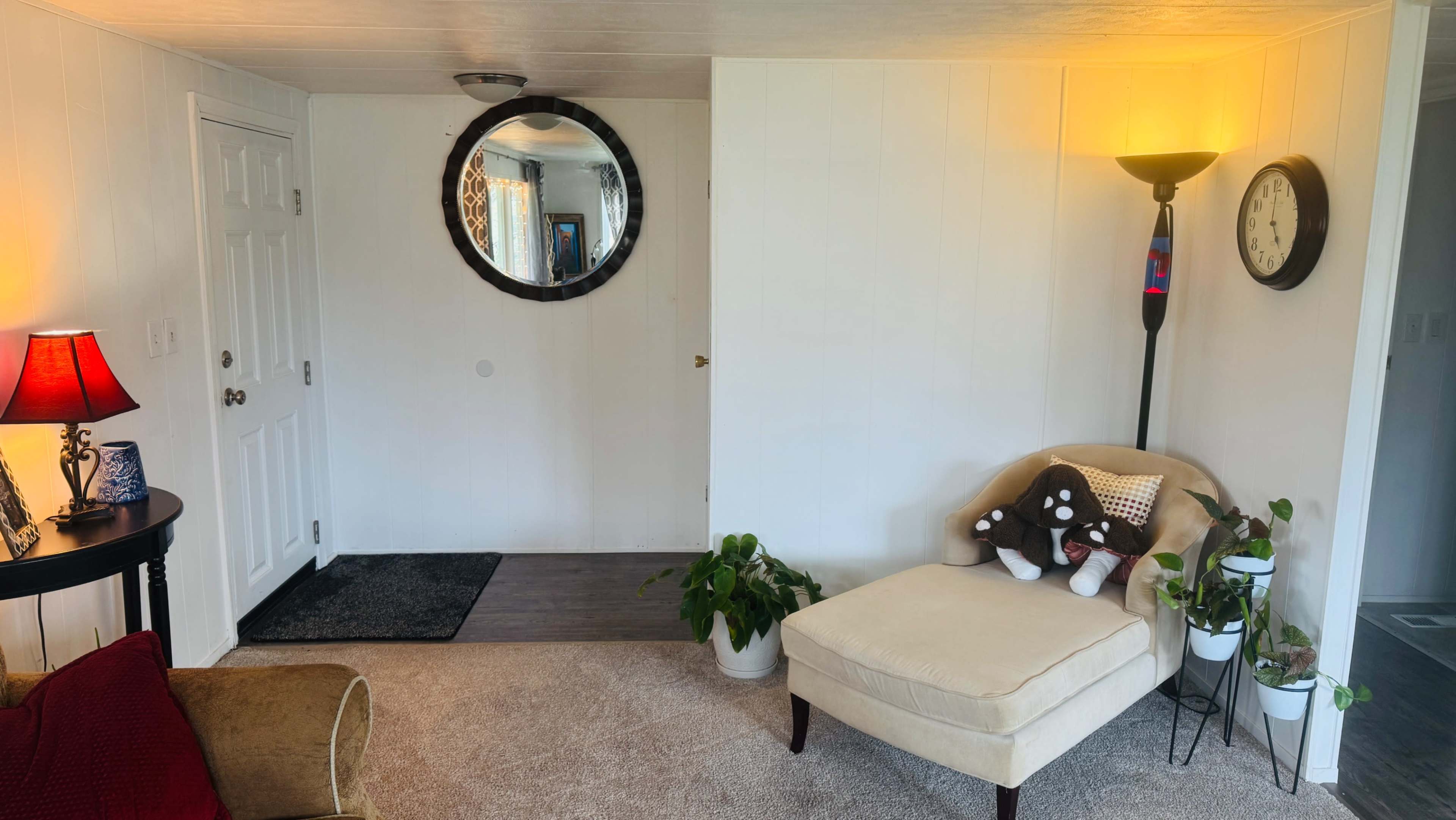 The image shows a brightly lit living space featuring a round mirror on the wall, a small chaise lounge with decorative pillows, a potted plant, a standing lamp, and an entrance door.