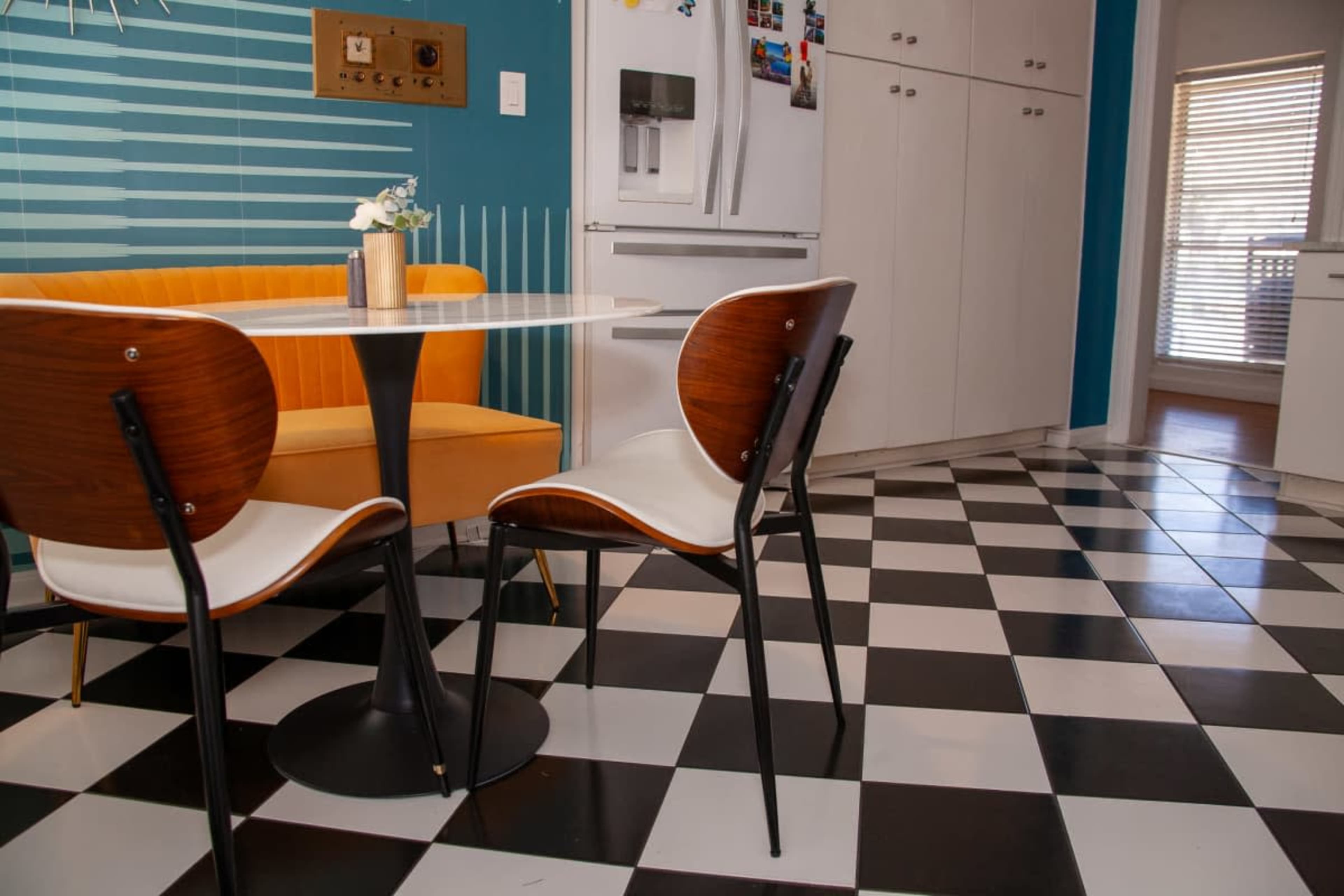 The image shows a kitchen with a round black and white checkered floor, a circular table with an orange banquette and two white-chairs, and a refrigerator against a bright blue wall.