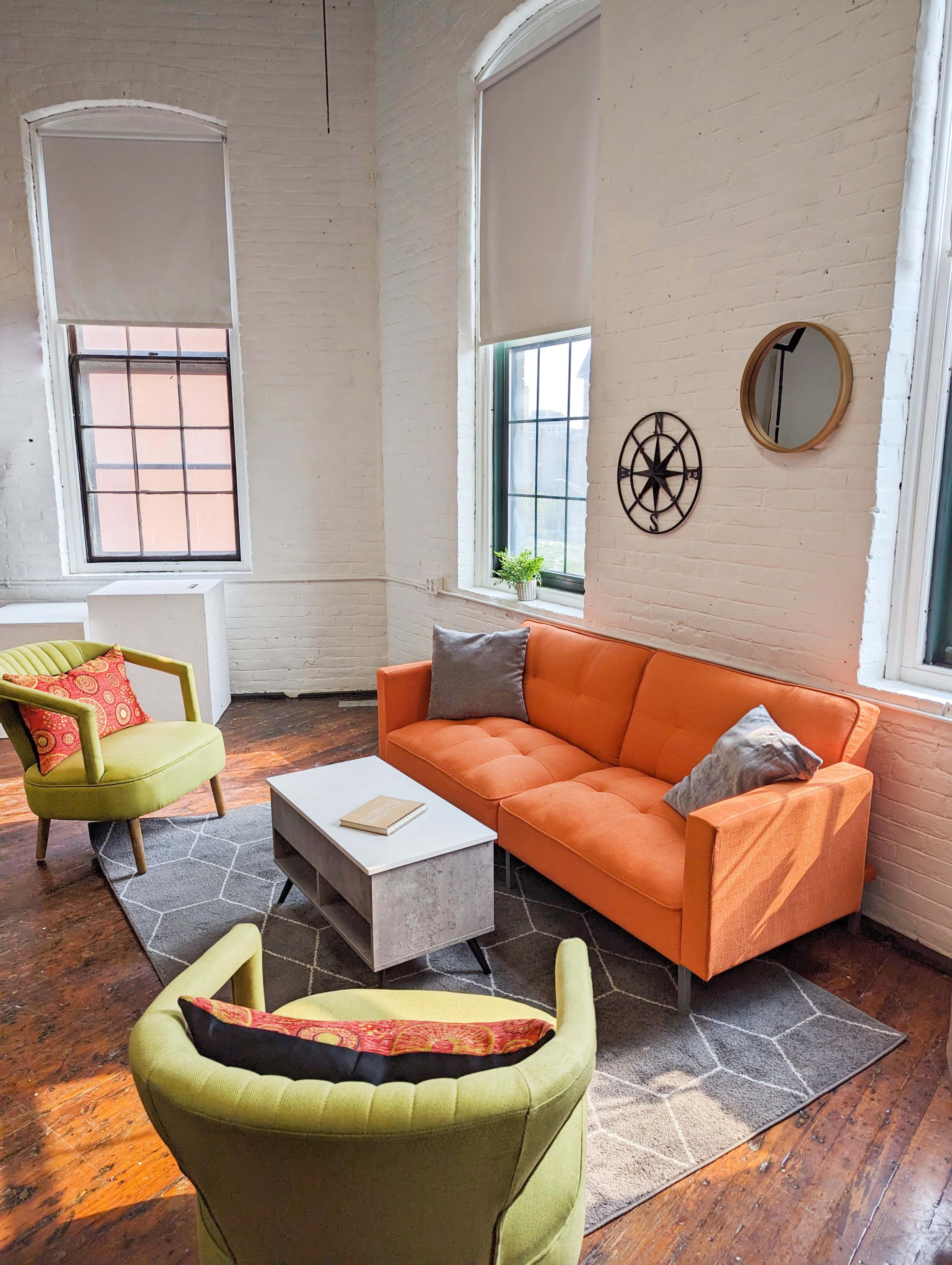 A bright, airy room featuring an orange sofa, two green chairs, a coffee table, and large windows with light streaming in.