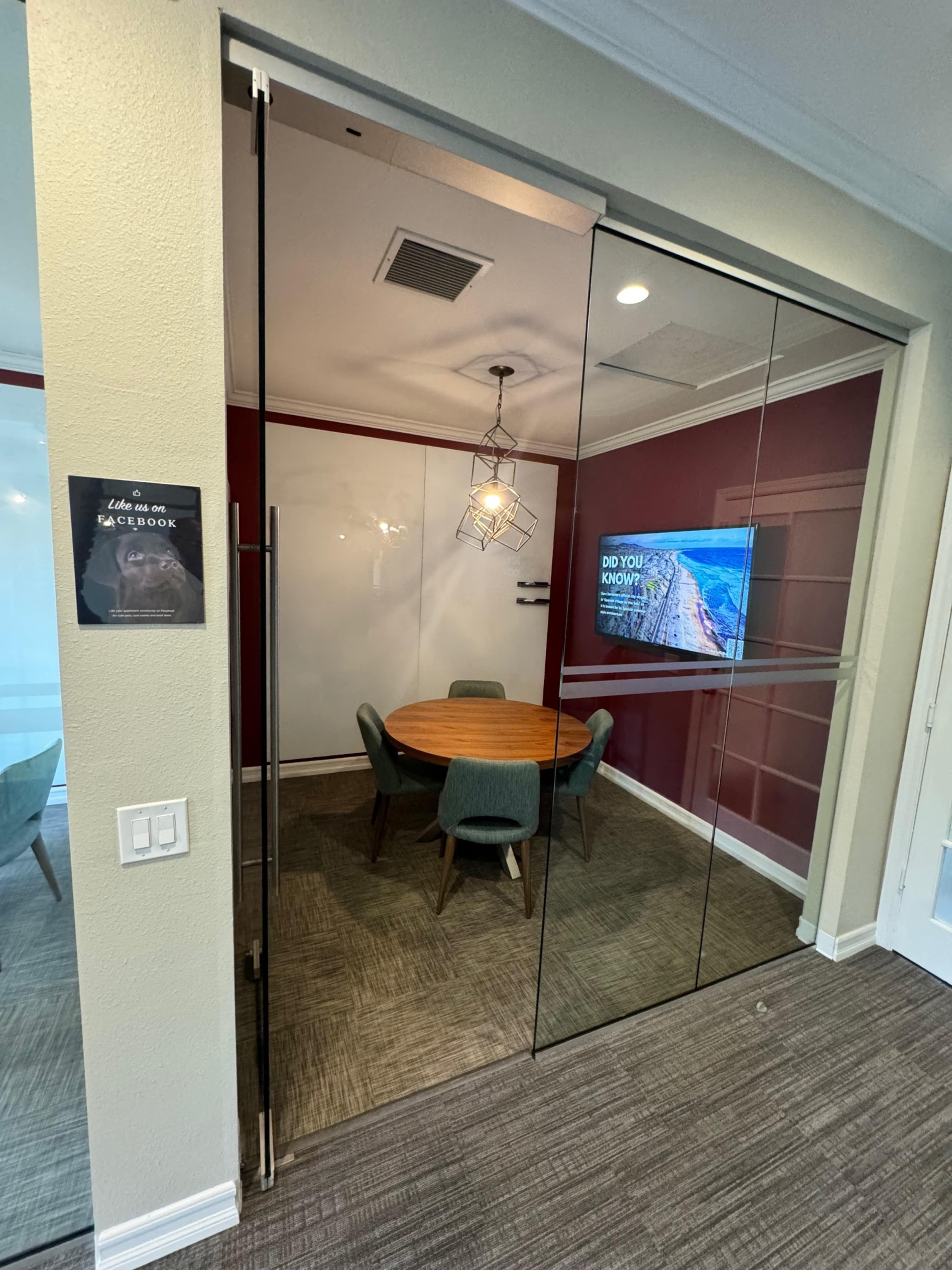 A conference room featuring a round wooden table surrounded by four green chairs, with a television mounted on the wall and large glass doors.