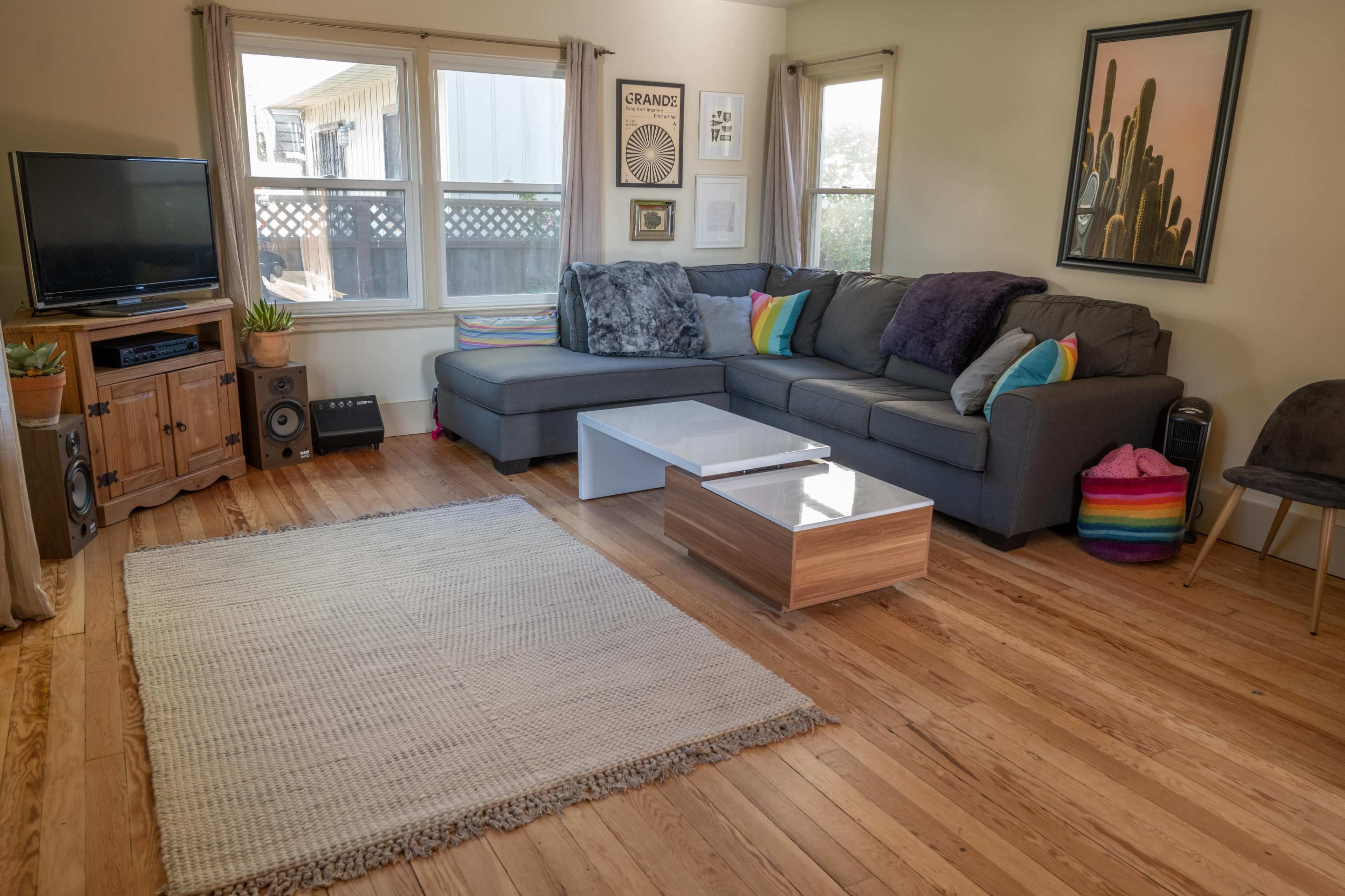 The living room features a gray sectional sofa, a wooden coffee table, a television, and a woven area rug on hardwood floors, with large windows allowing natural light.