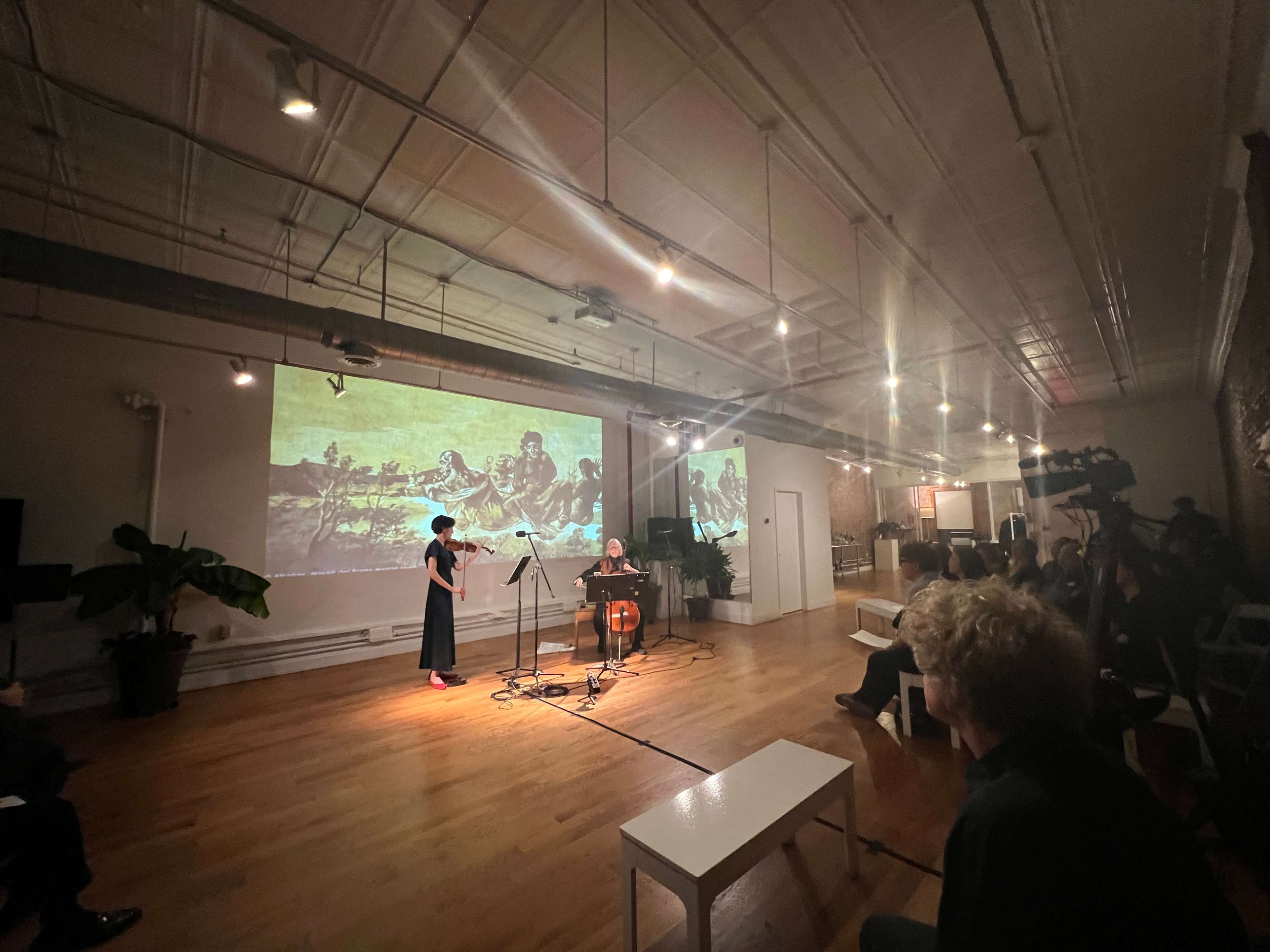 A performer in a black dress stands beside a cellist in a spacious room while an artistic projection fills the wall behind them.