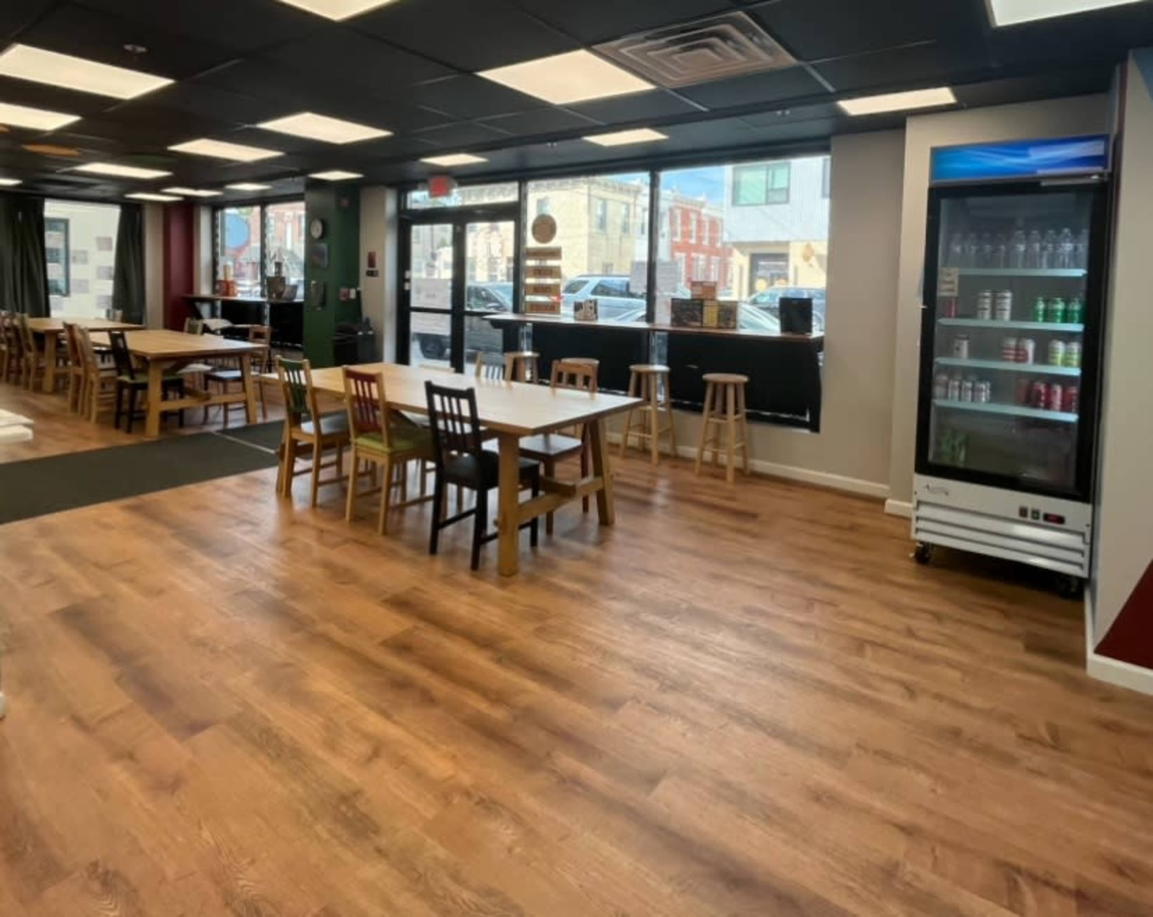 The image shows a spacious restaurant interior with wooden tables and chairs, large windows, and a beverage cooler displaying drinks.
