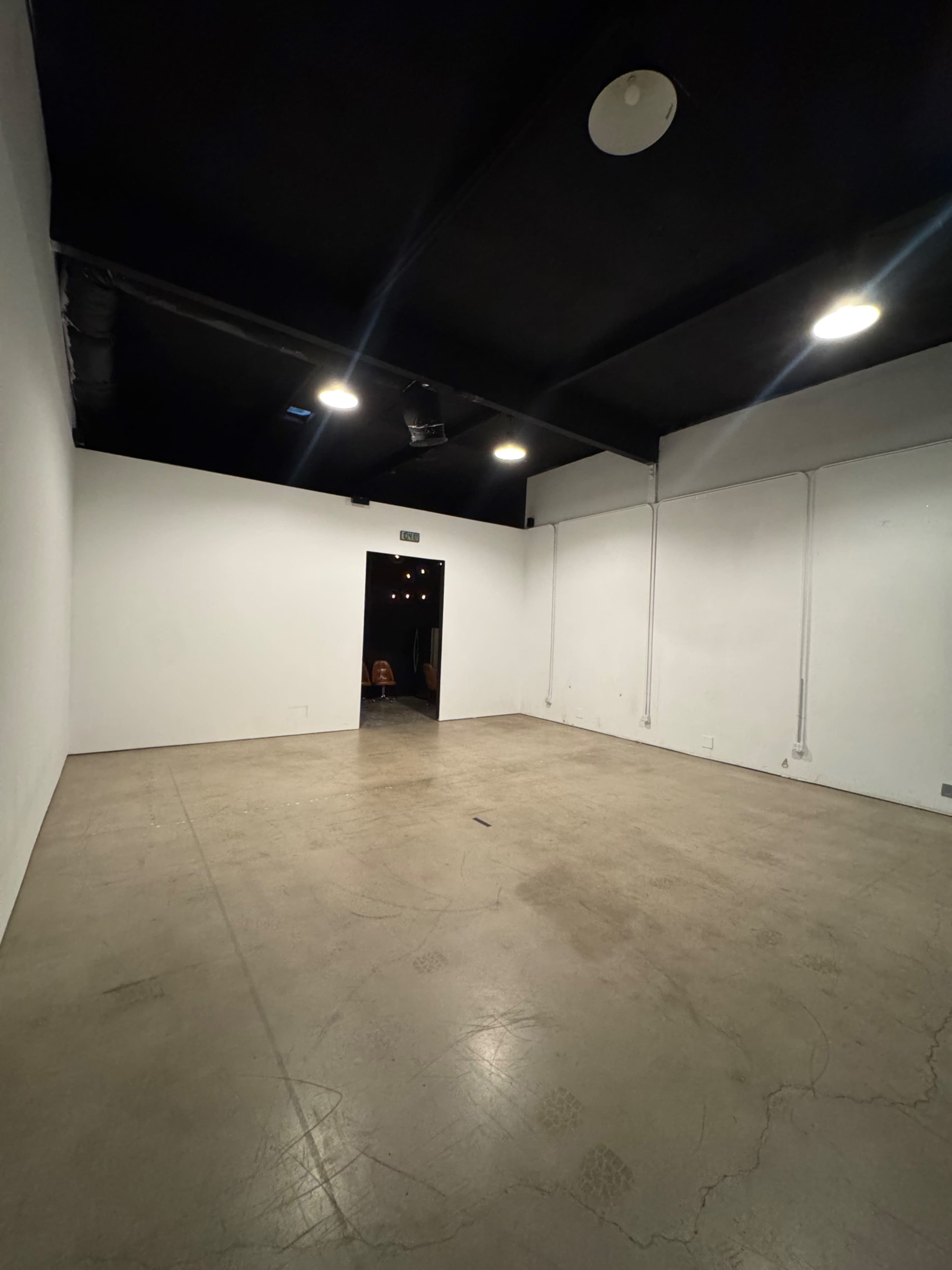 The image shows a sparsely furnished room with a concrete floor, white walls, and a doorway leading to another area.