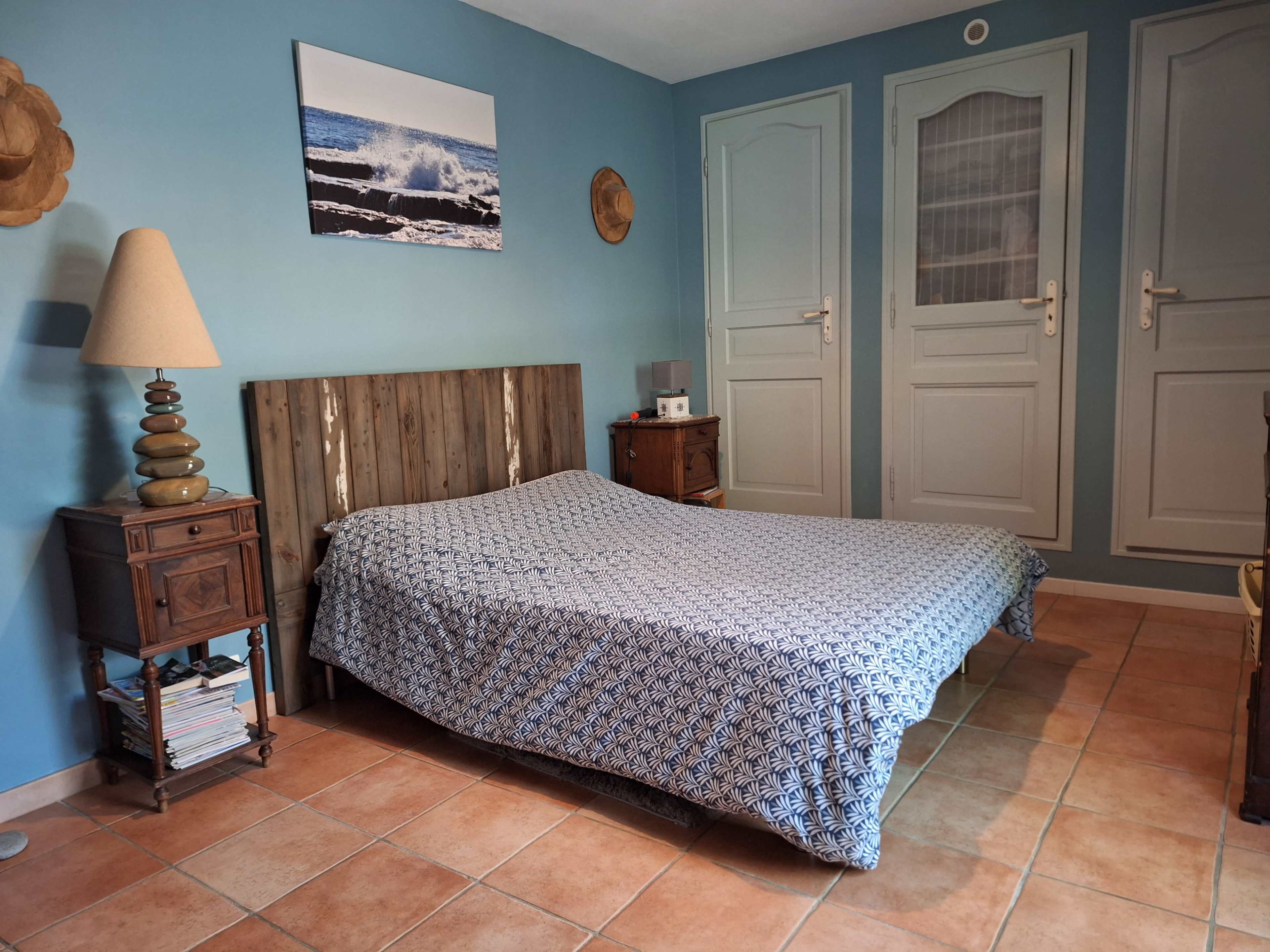 The image shows a bedroom with a large bed covered by a patterned blanket, a wooden headboard, a lamp beside the bed, and two doors in the background.