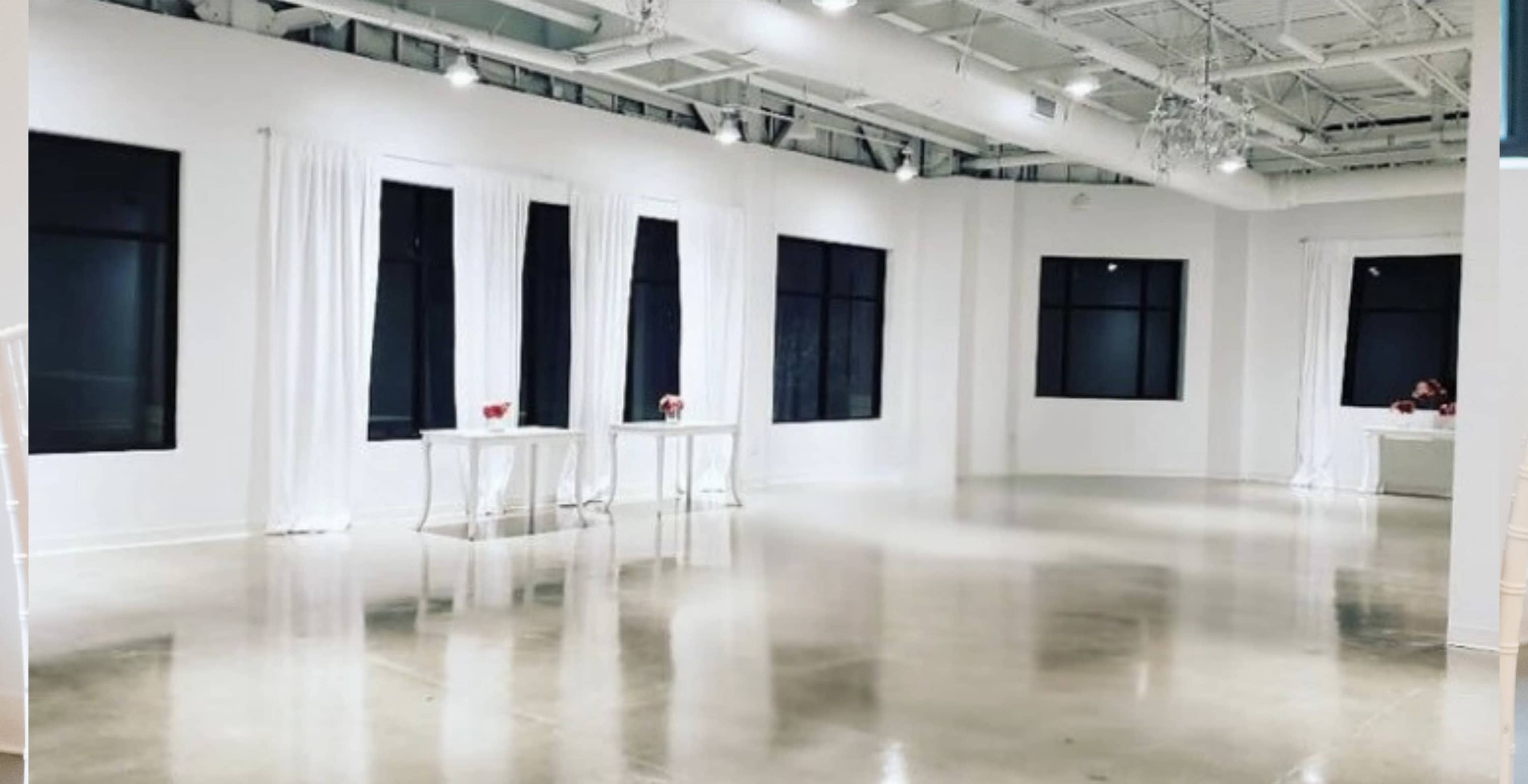 The image shows a spacious, empty room with white walls, large windows, and polished concrete floors, featuring two small white tables decorated with red flowers.