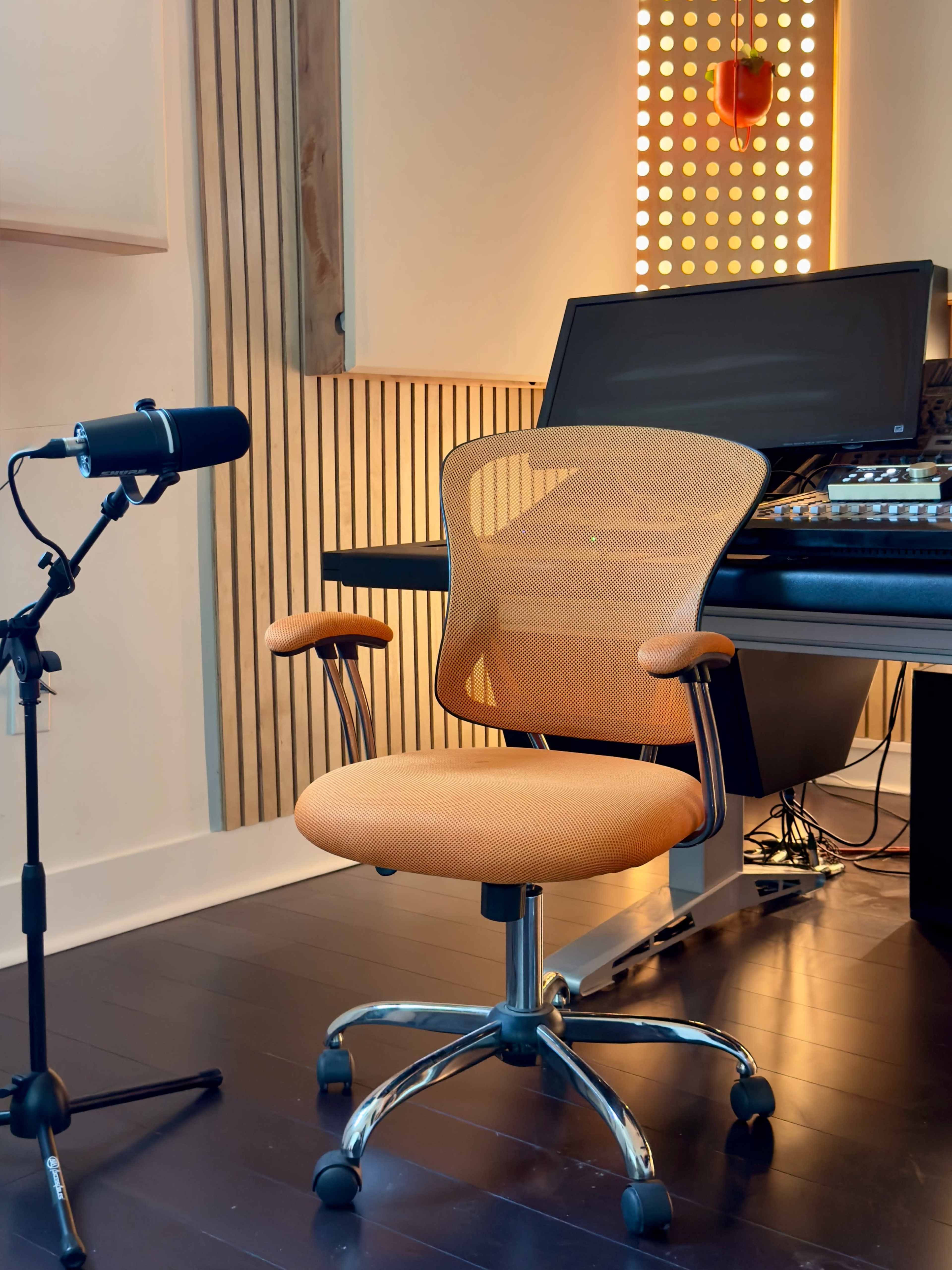 The image shows an orange office chair positioned beside a microphone, with a music production workstation in the background.