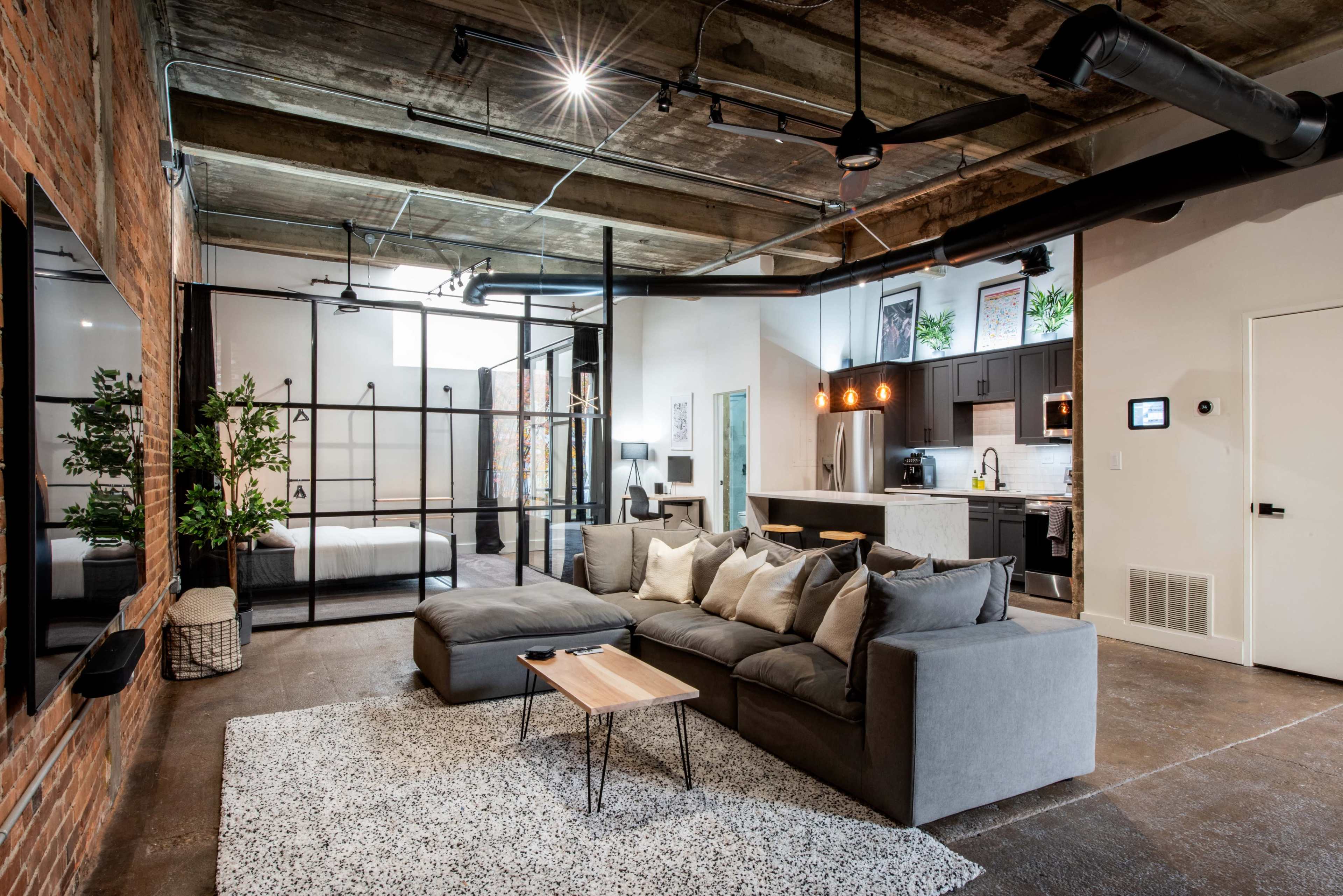 A modern open-concept living space with a sectional sofa, a small wooden coffee table, a kitchen area, and a glass-walled bedroom at the back.