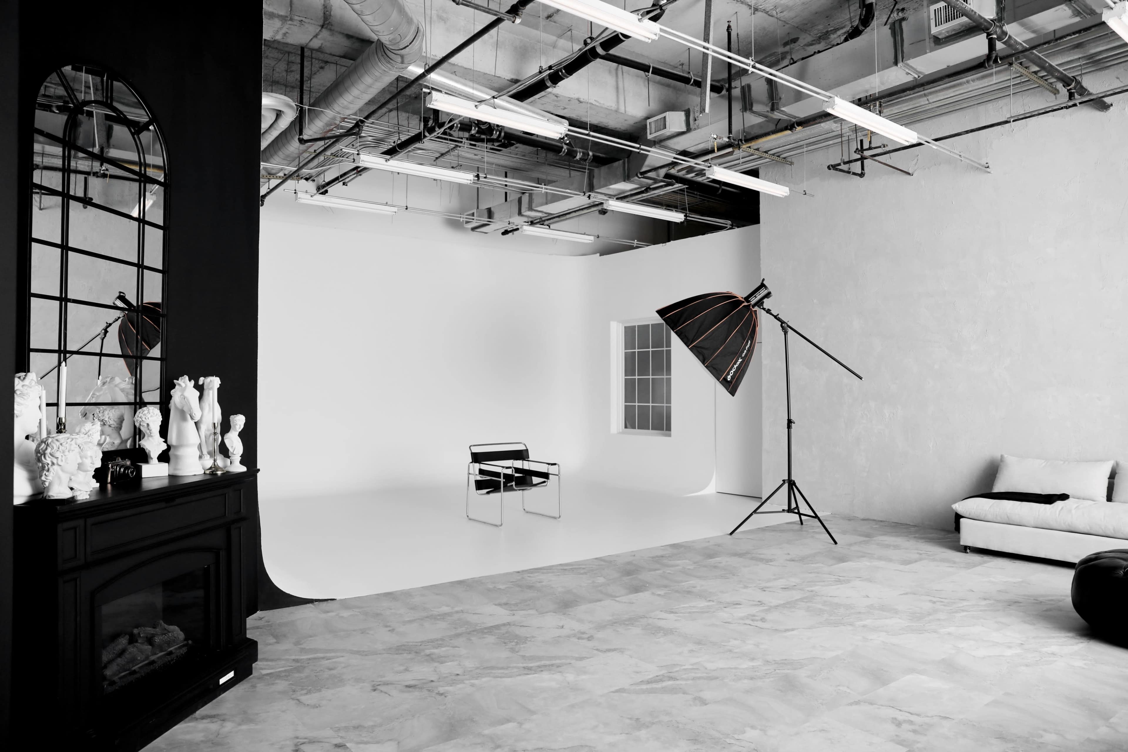 A minimalist photography studio featuring a black-and-white color scheme, with a large backdrop, a chair, a softbox light, and a decorative mirror on one side.