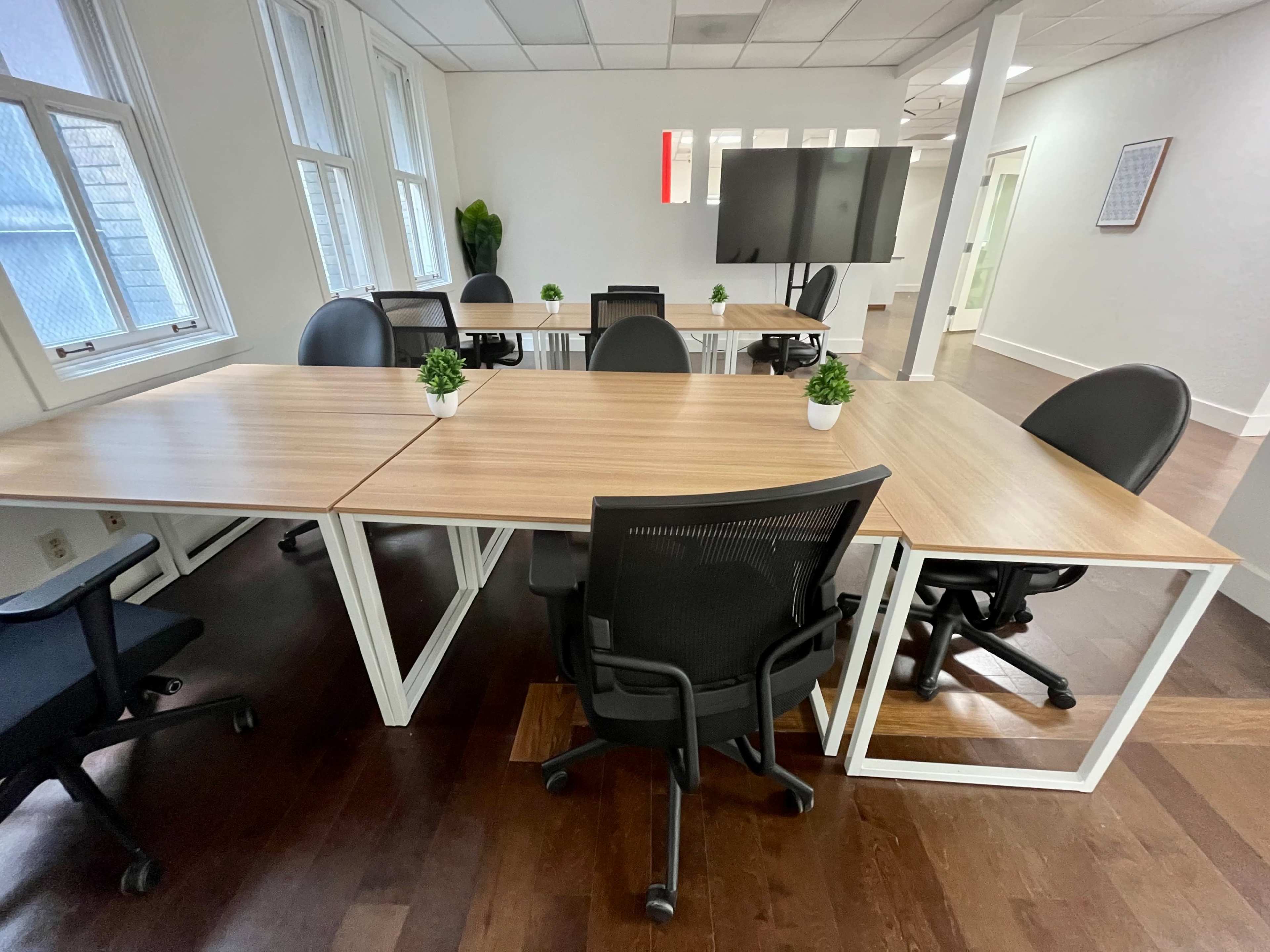 A modern office space features several tables with black chairs and small potted plants, surrounded by large windows and a wall-mounted screen.