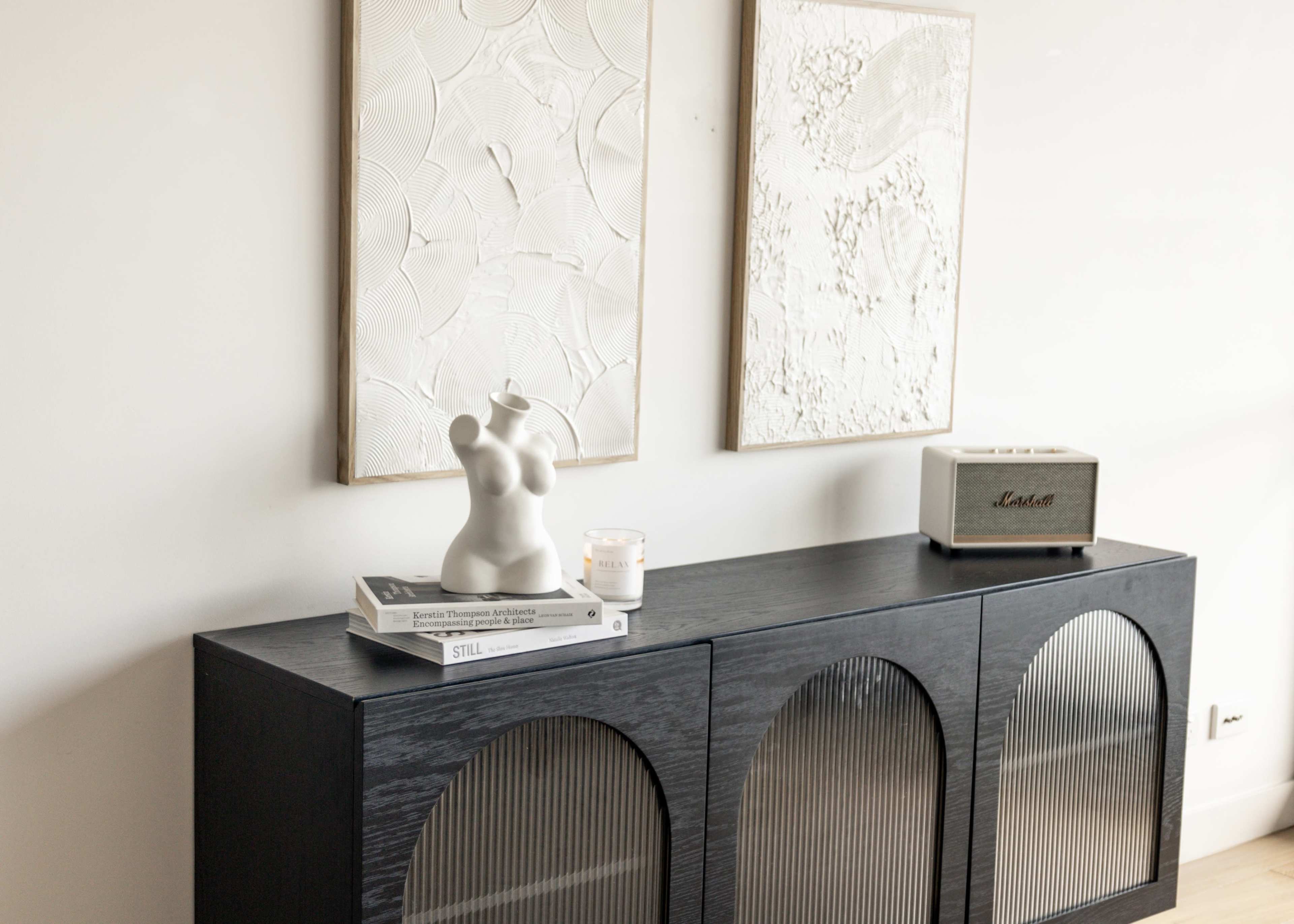 A black wooden console table holds books, a candle, a sculptural piece, and a speaker, with two framed textured artworks displayed above it.