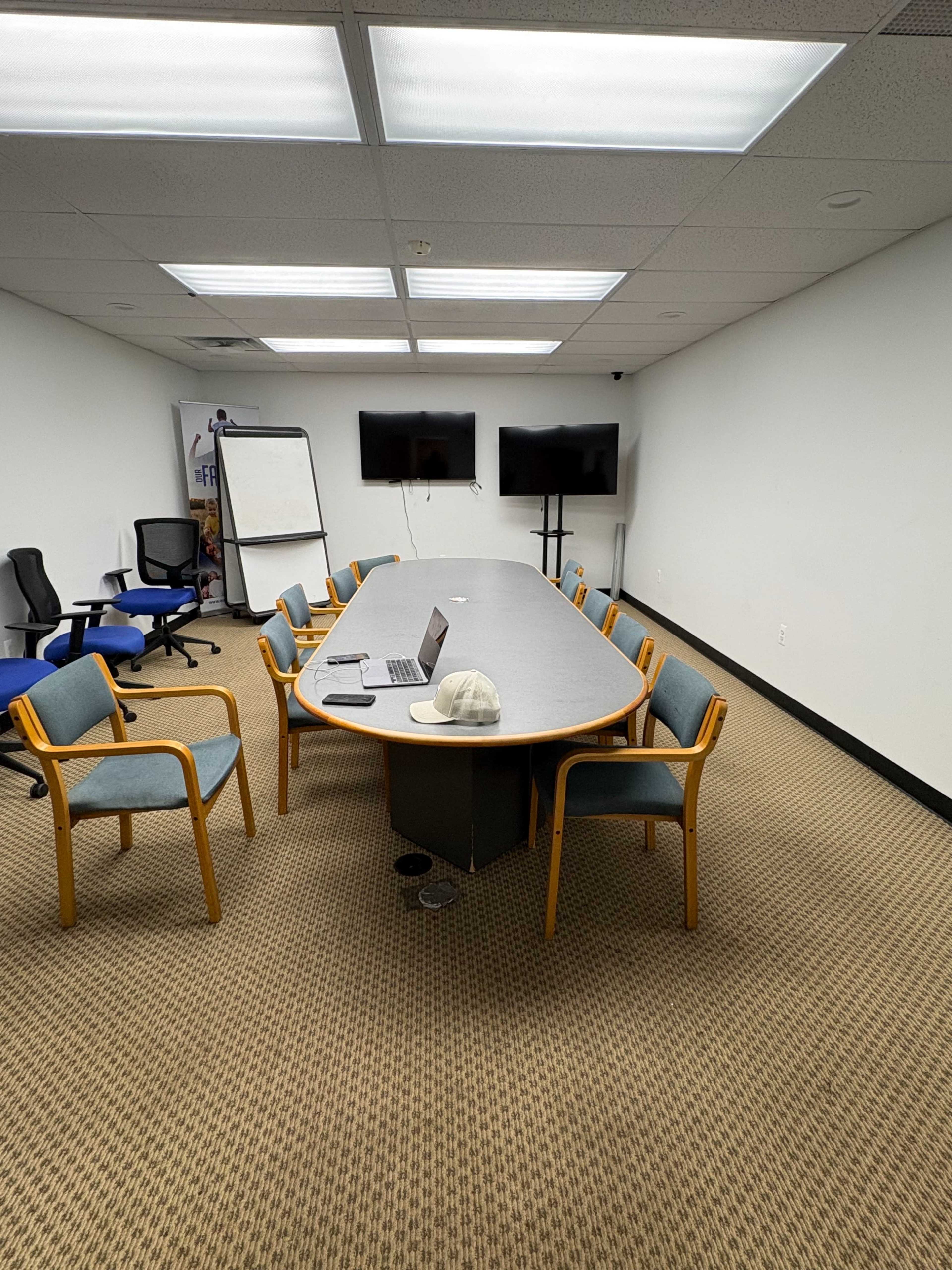 The image shows a conference room with a long table surrounded by chairs, featuring a laptop and a cap on the table, along with two monitors on the wall.