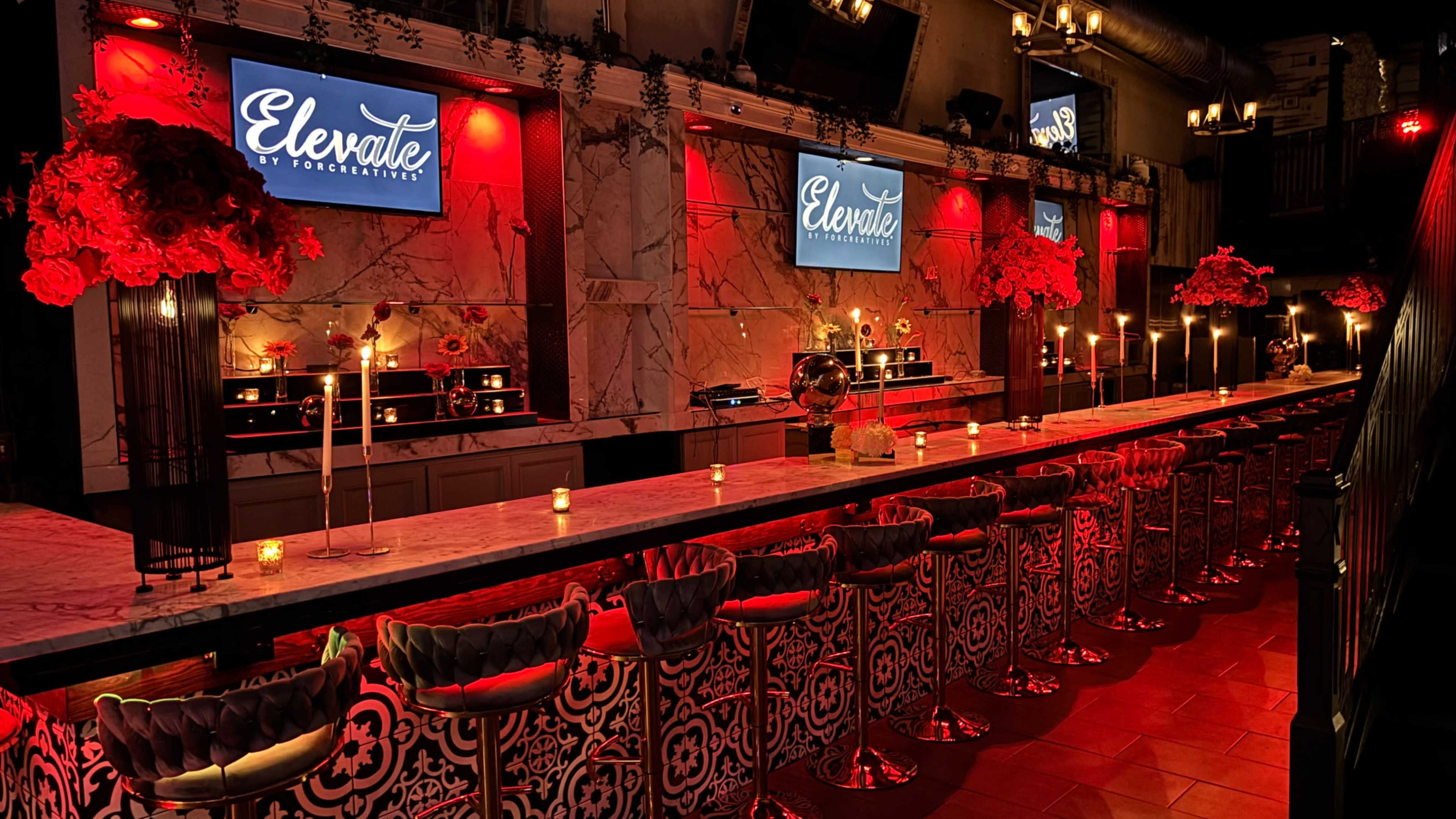 A stylish bar area features a long marble counter with plush stools, illuminated by red lights and decorated with flowers and candles.