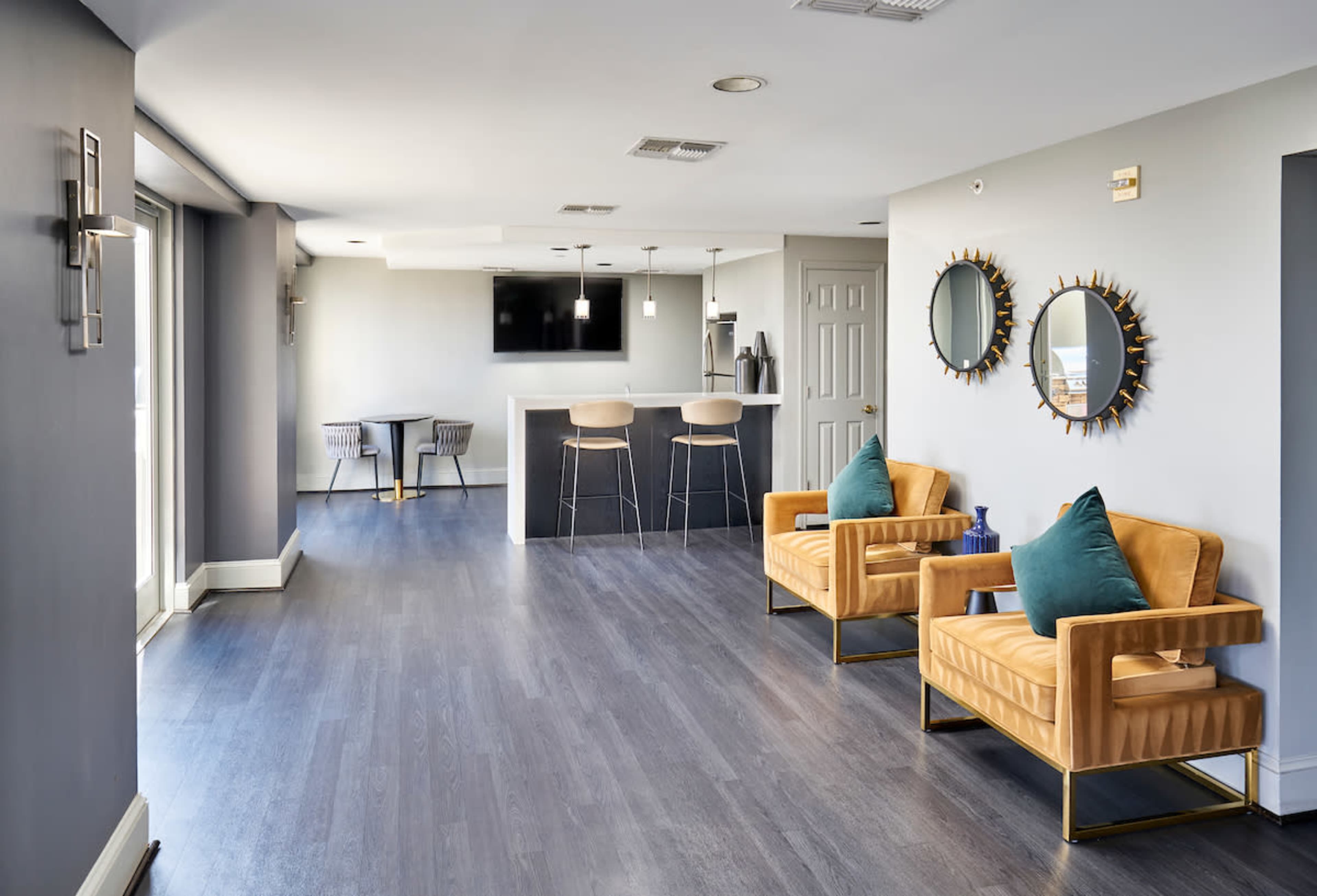 A modern lobby area with two yellow armchairs, a small table, and a bar counter, featuring mirrors on the walls and a TV mounted above.