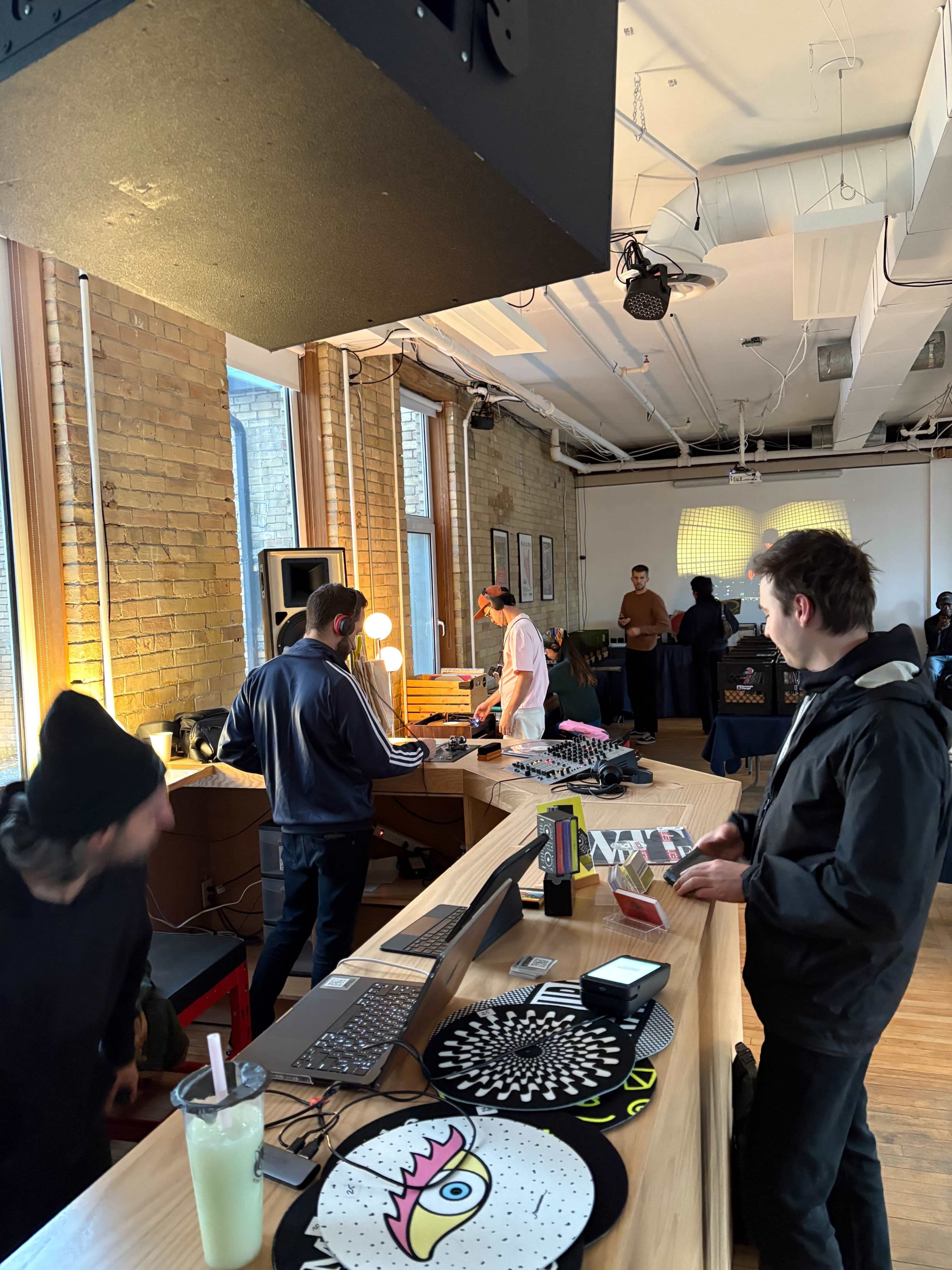 A group of people are gathered inside a well-lit room with wooden floors, engaging with music equipment and DJing setup on a long wooden table.