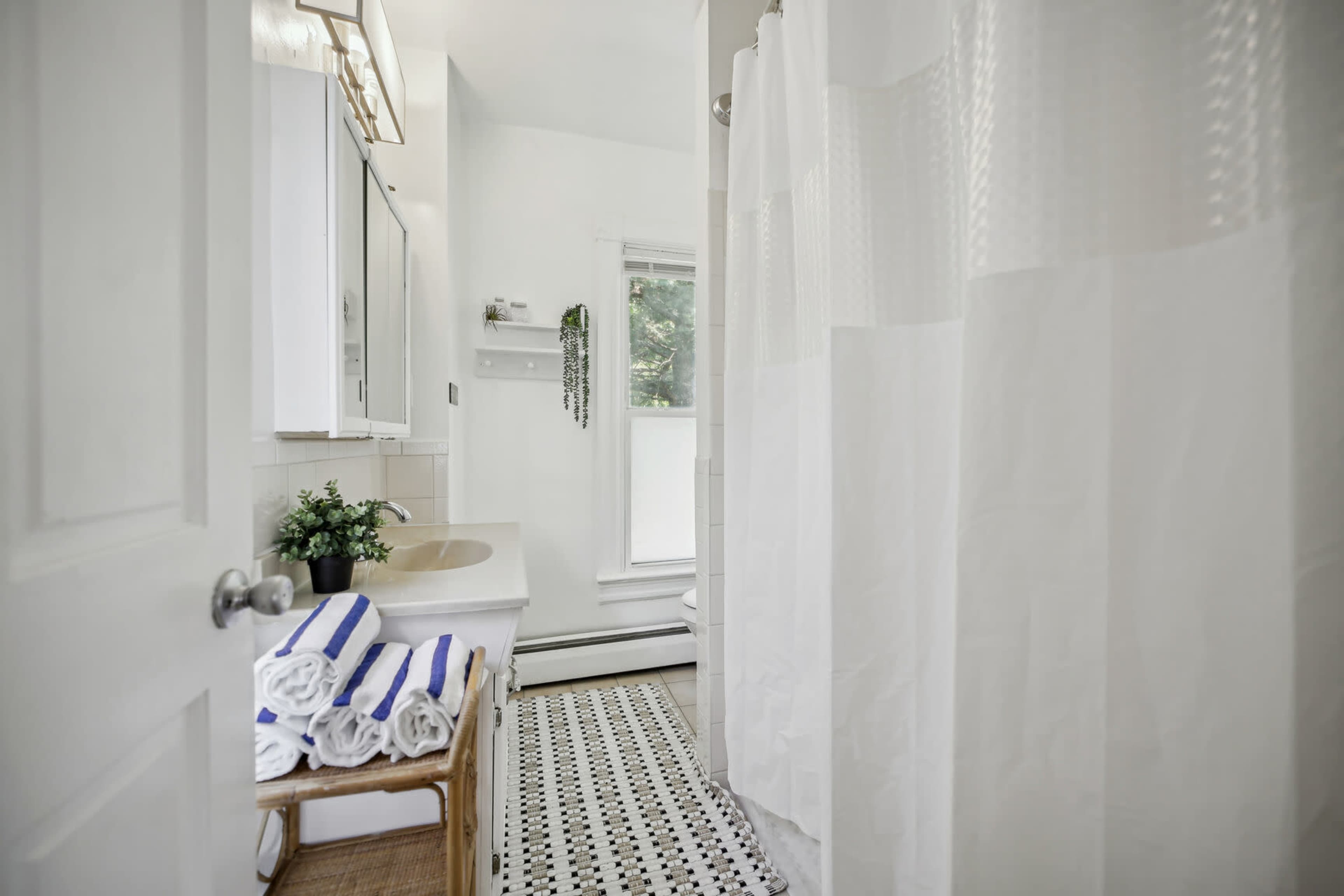 A bright bathroom features a walk-in shower with a white curtain, a small vanity, and neatly stacked towels on a wooden rack.