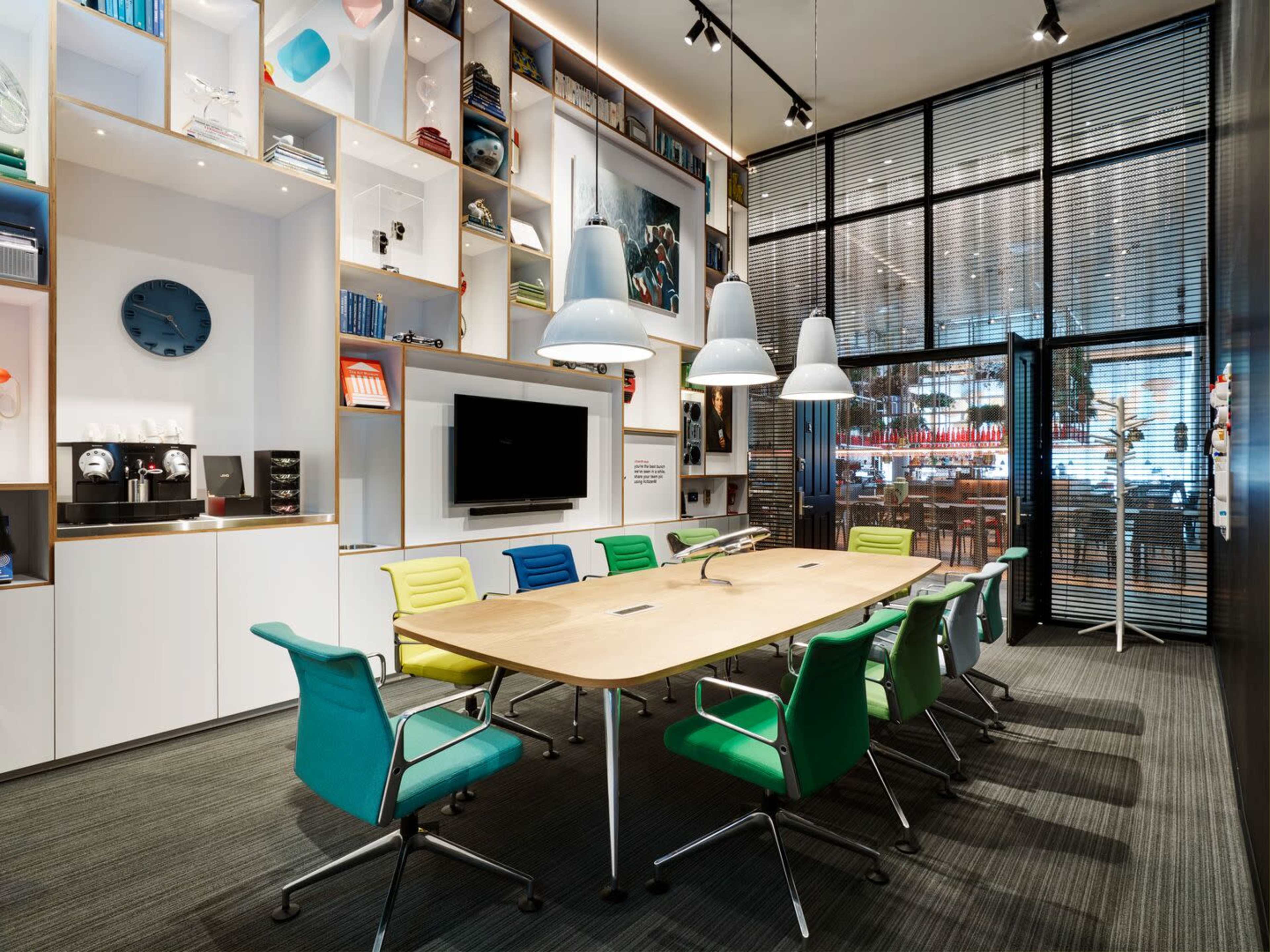 A modern conference room featuring a large wooden table surrounded by colorful chairs, a wall of shelves filled with books and decor, and glass doors leading to an adjacent area.