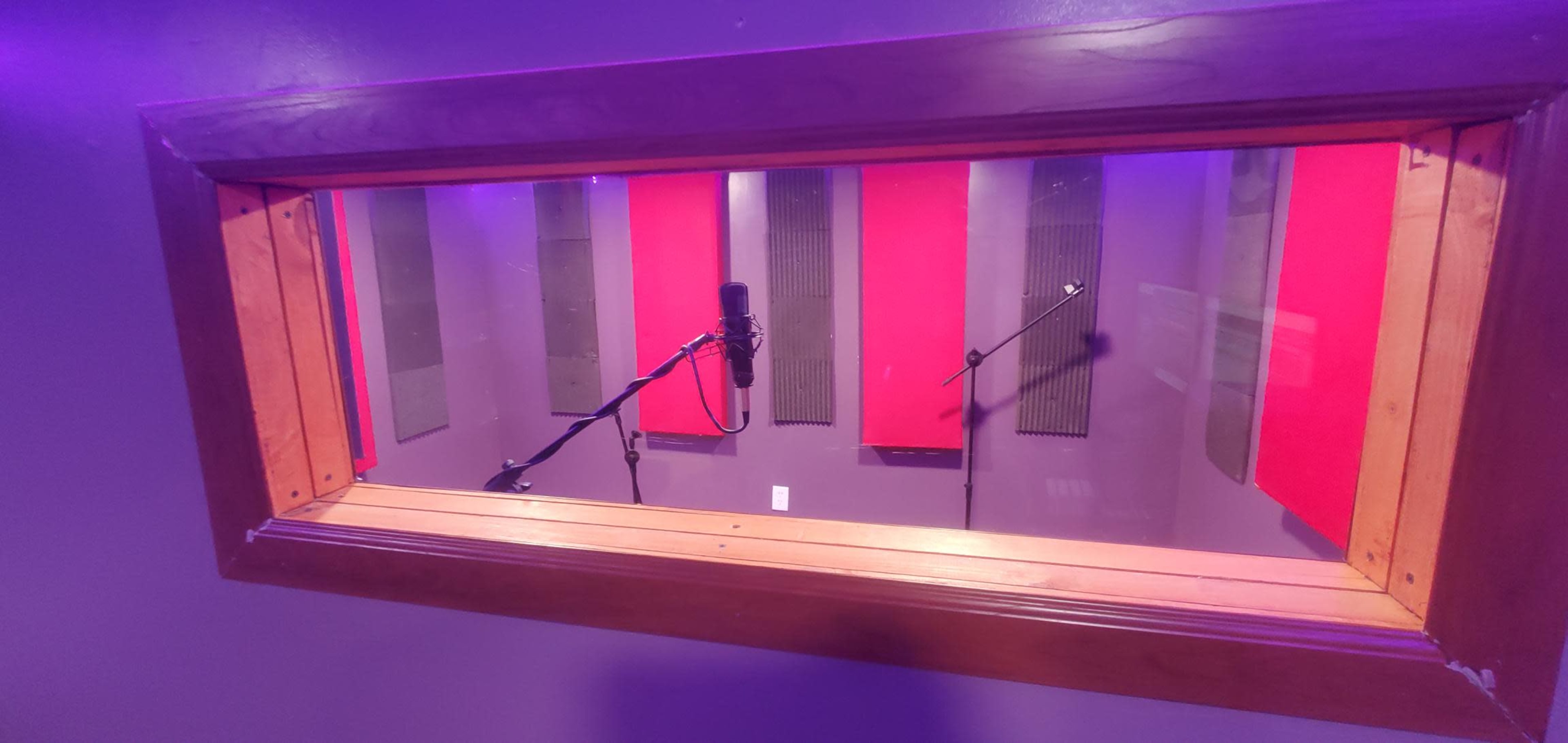 A recording studio booth viewed through a window, featuring microphones set up amidst soundproofing panels.