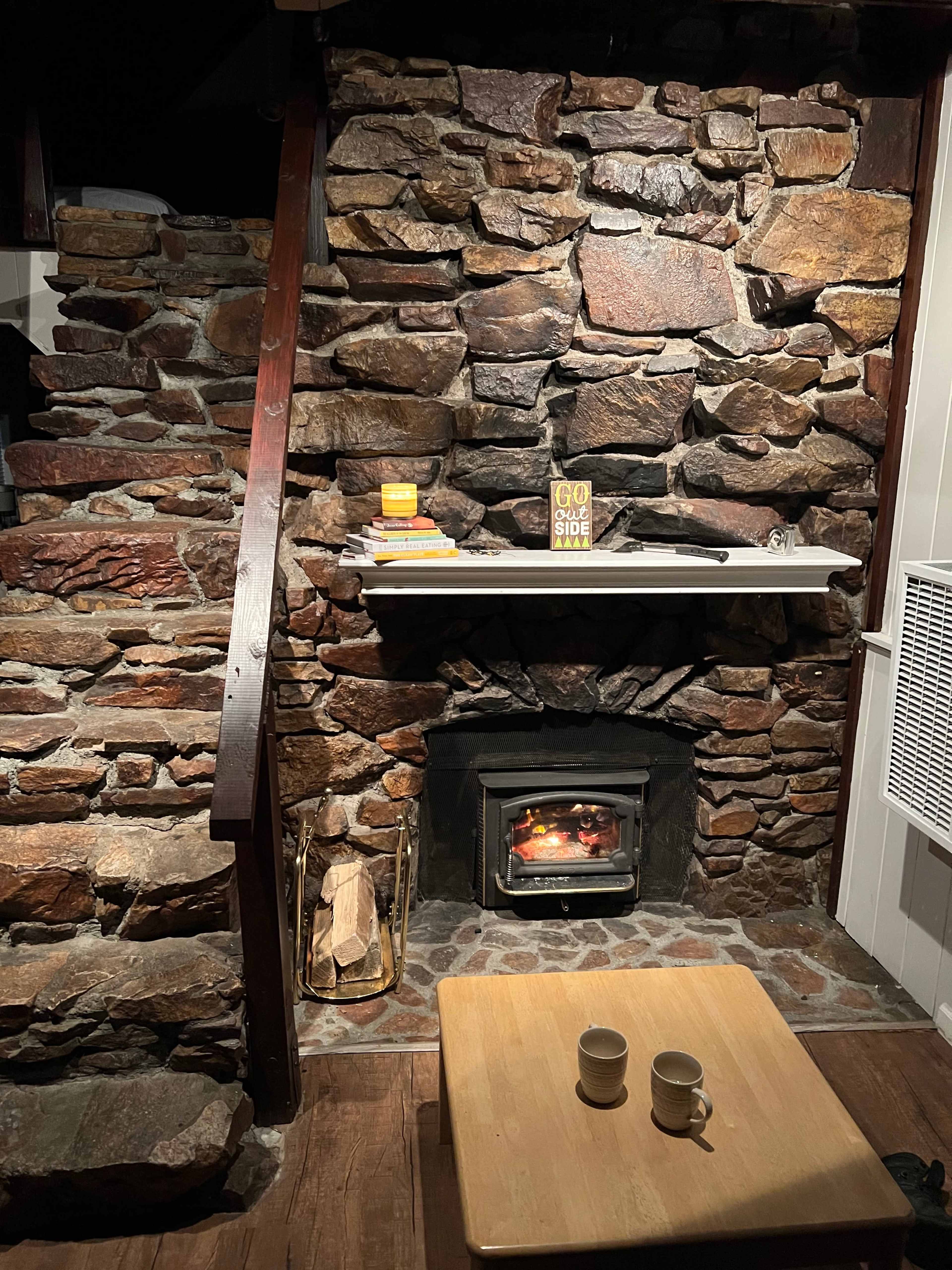 A stone fireplace with a wood-burning stove is set against a wall, with a small wooden table and mugs in front, and a staircase leading up to the left.