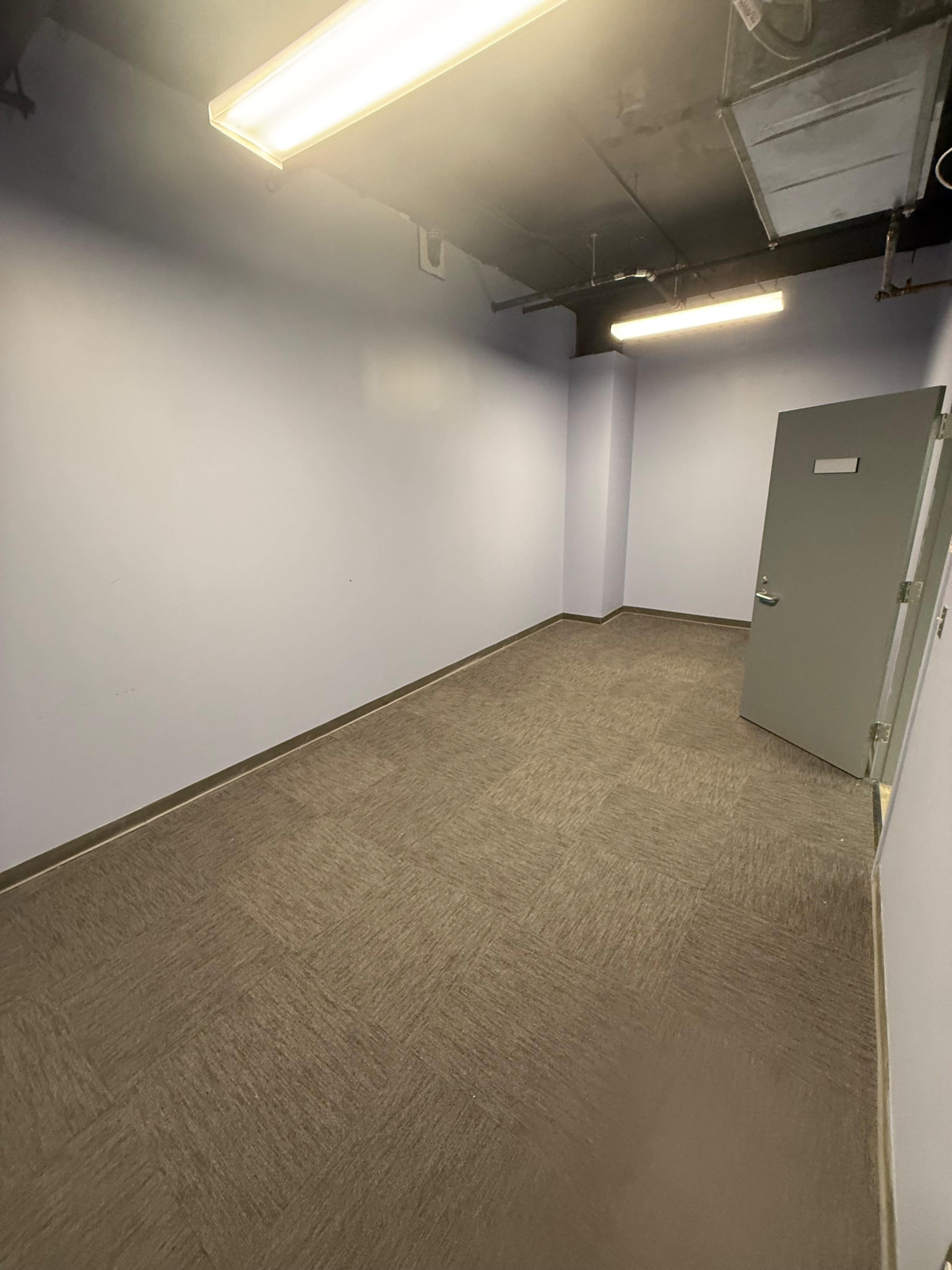 The image shows an empty room with light purple walls, gray carpet, and a gray door on one side.