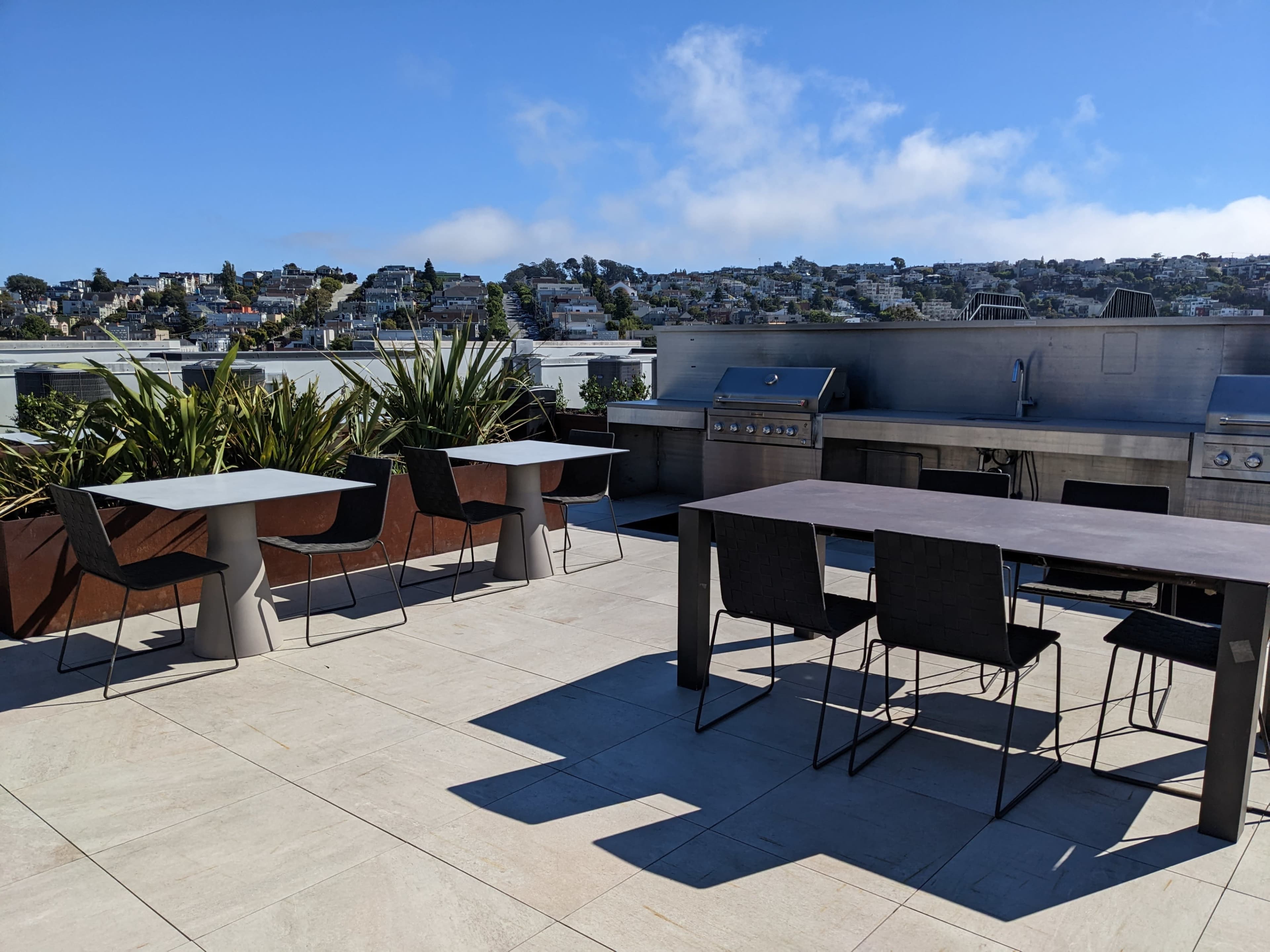 A rooftop terrace features a dining table and seating, with a grill station and a view of a hillside neighborhood.