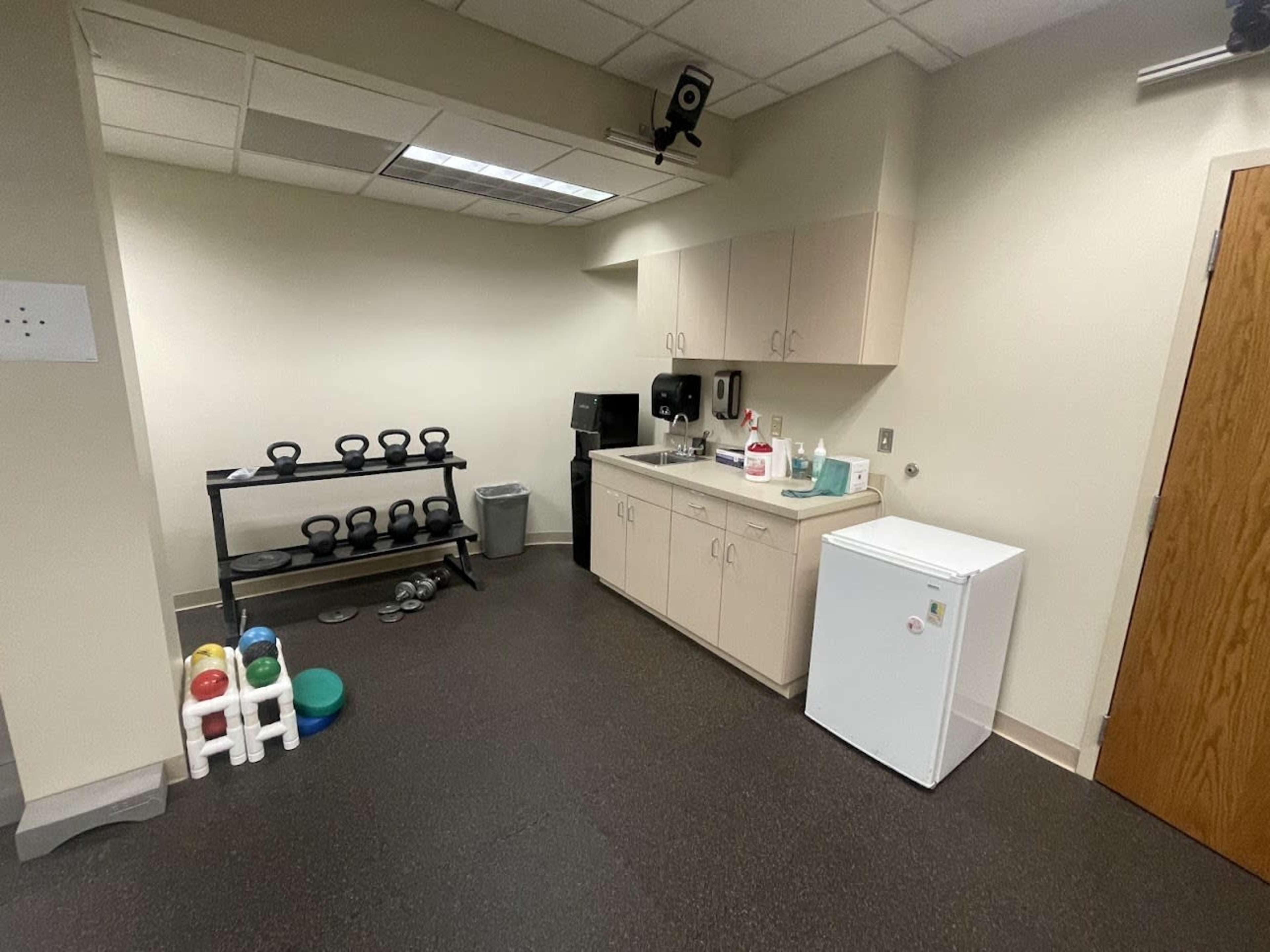 A small kitchenette area with a sink, cabinets, a refrigerator, and a shelf holding kettlebells, alongside a trash can and some exercise equipment.