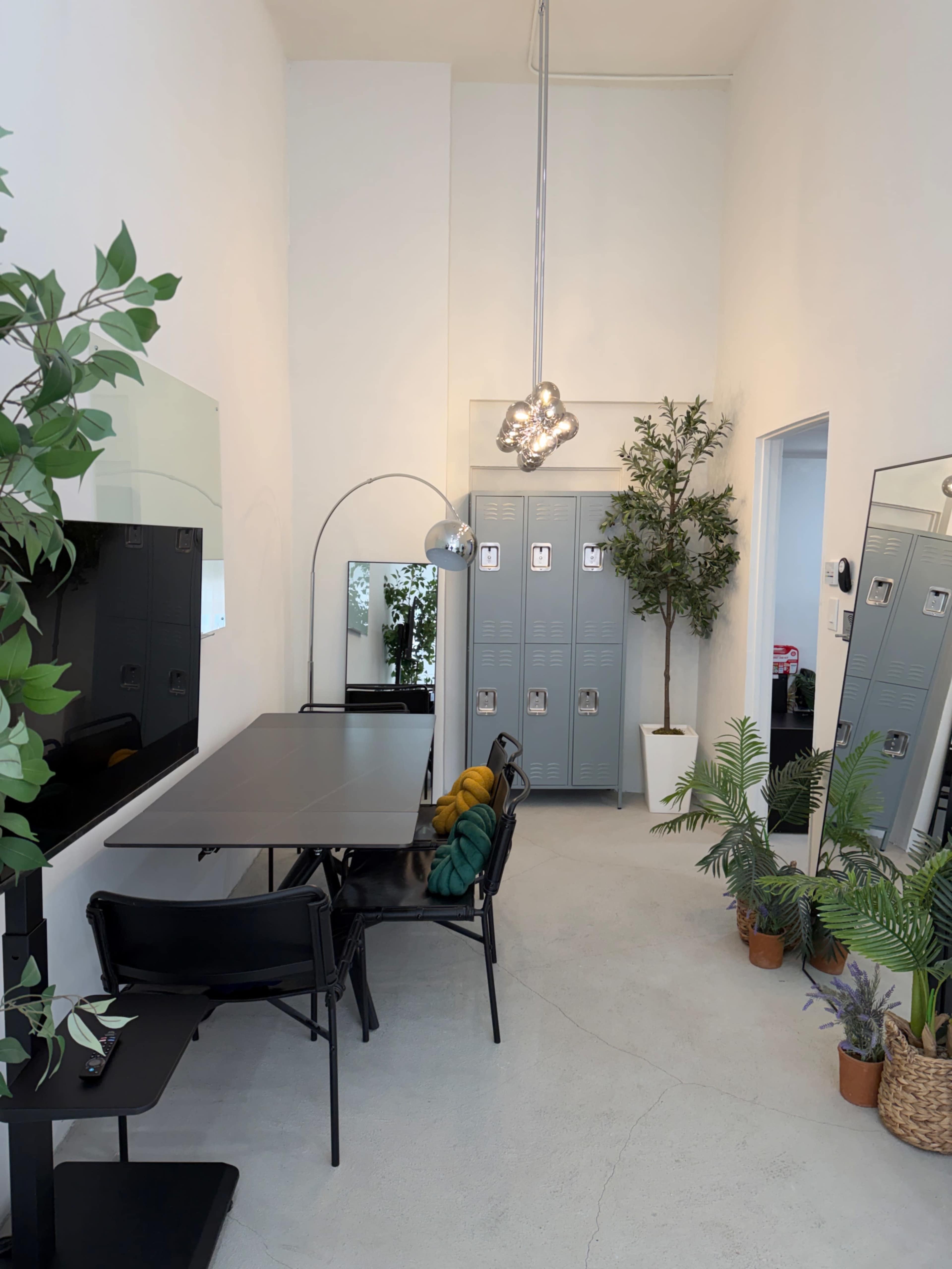 The image shows a narrow, modern workspace with a table and chairs, greenery, lockers, and a large mirror.