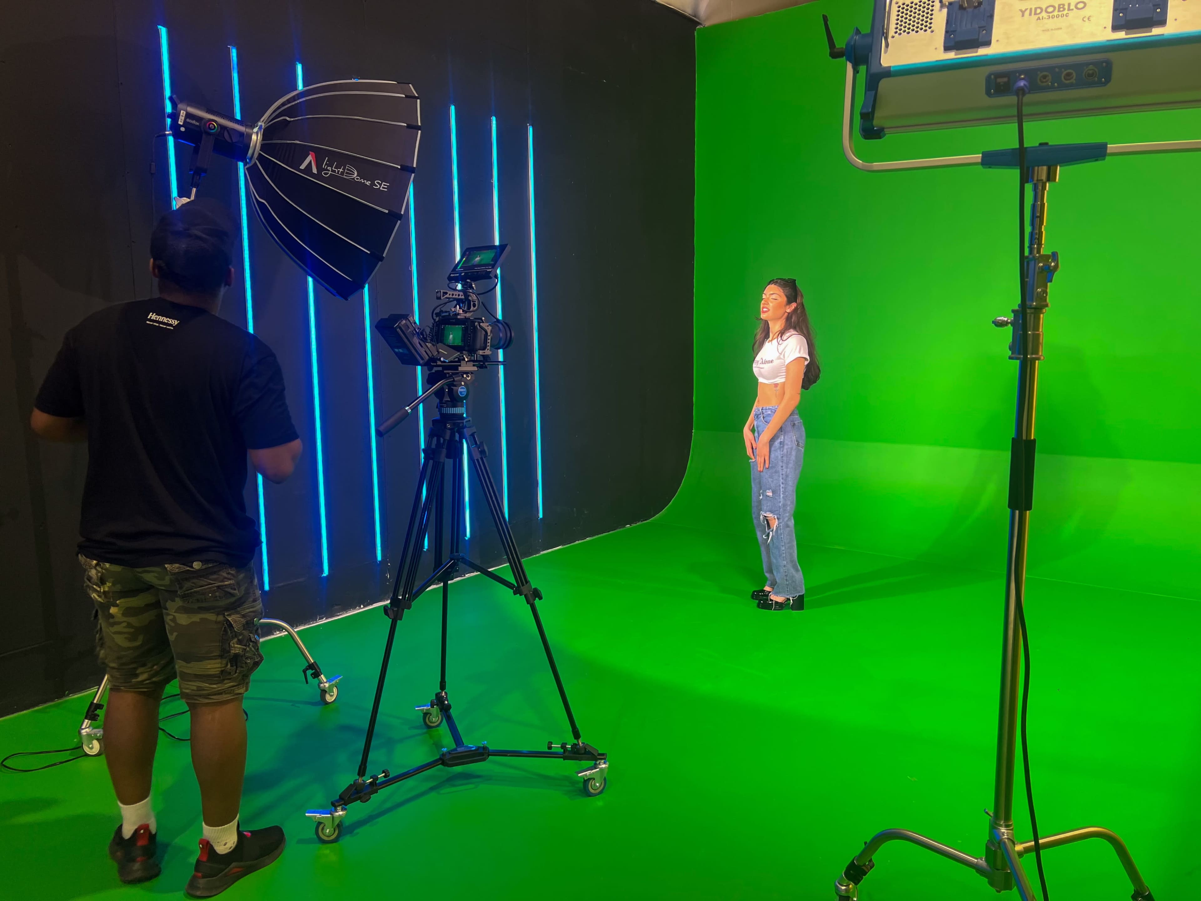 A person stands on a green screen set while a cameraman prepares to record with lighting equipment in the background.