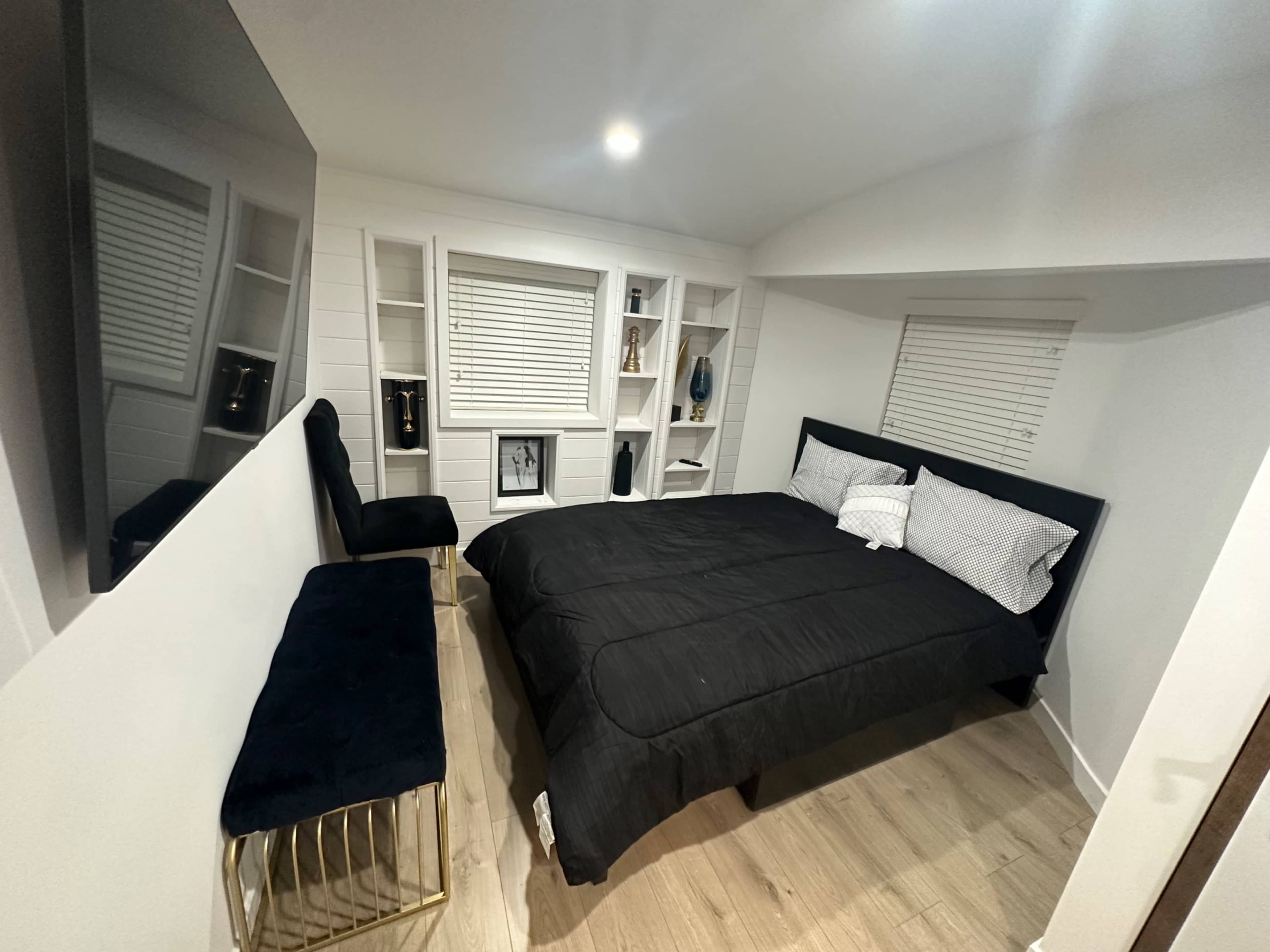 A modern bedroom featuring a black bed with white linens, a gold accent bench, and built-in shelving against a white wall.