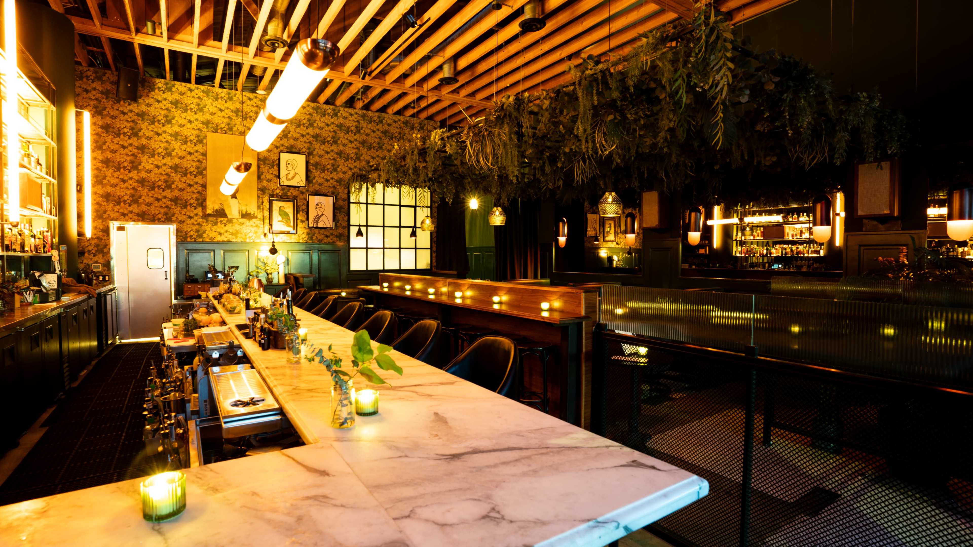 The image shows a dimly lit bar with a marble countertop, wooden beams overhead, and greenery adorning the ceiling.