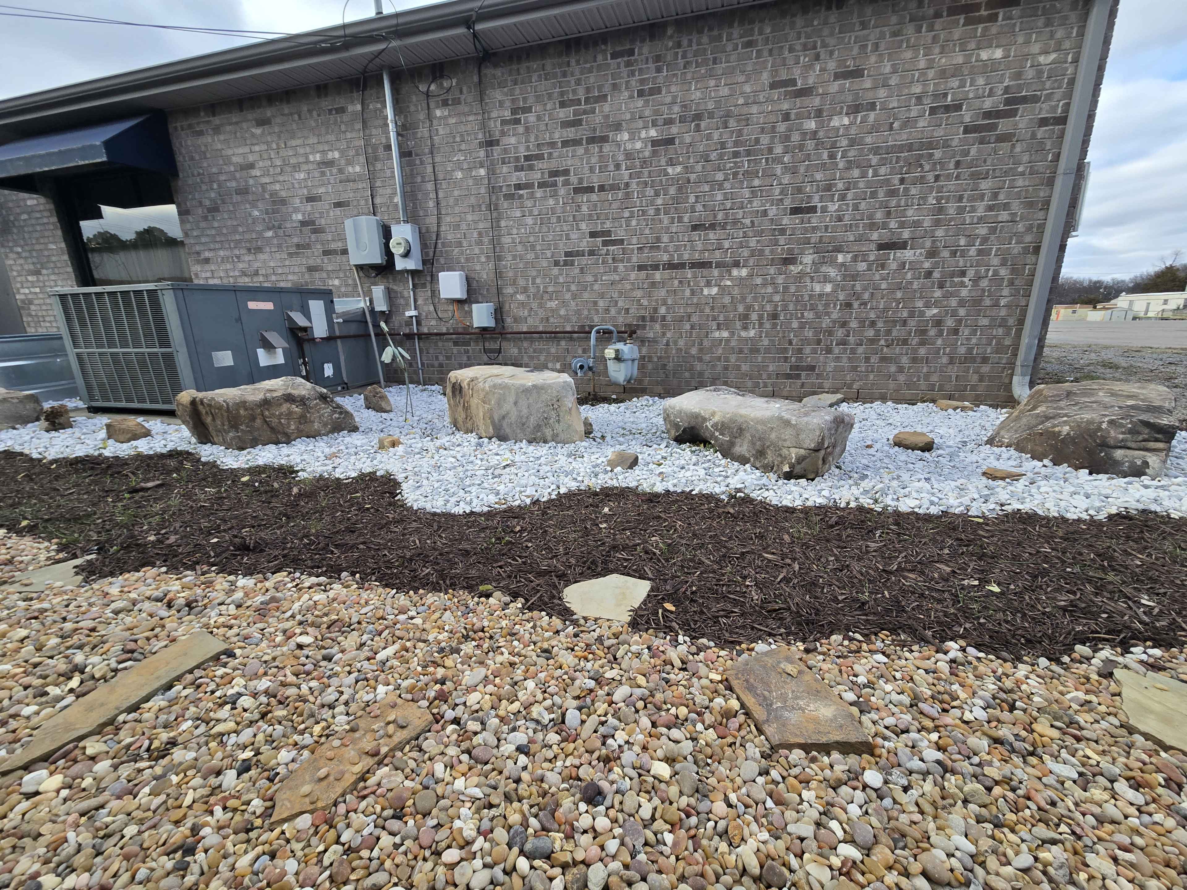 The image shows a landscaped area featuring large rocks, white gravel, and a bed of mulch arranged alongside a brick building.