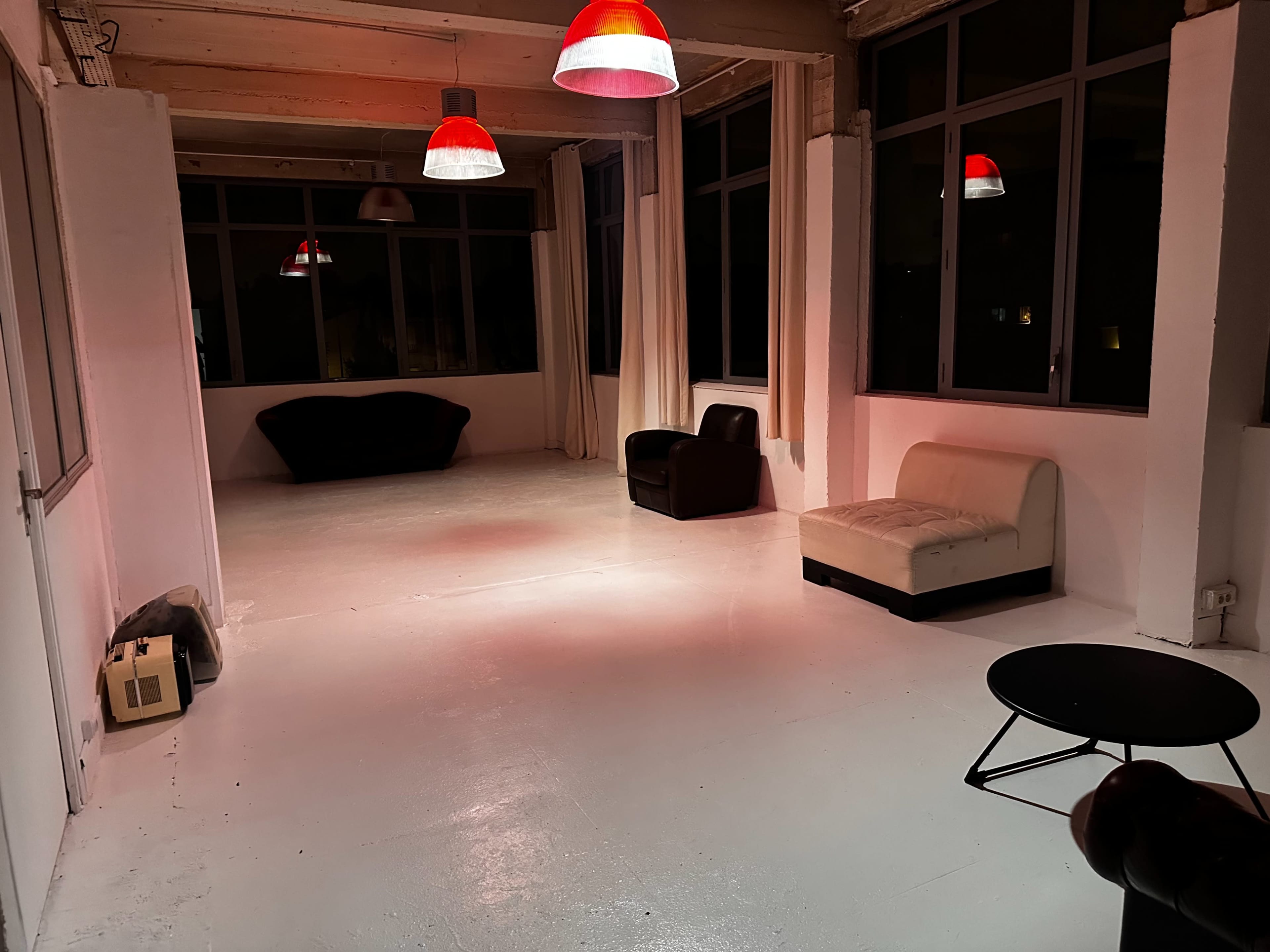 The image shows a spacious, minimally furnished room with two chairs and a small table, illuminated by red pendant lights.