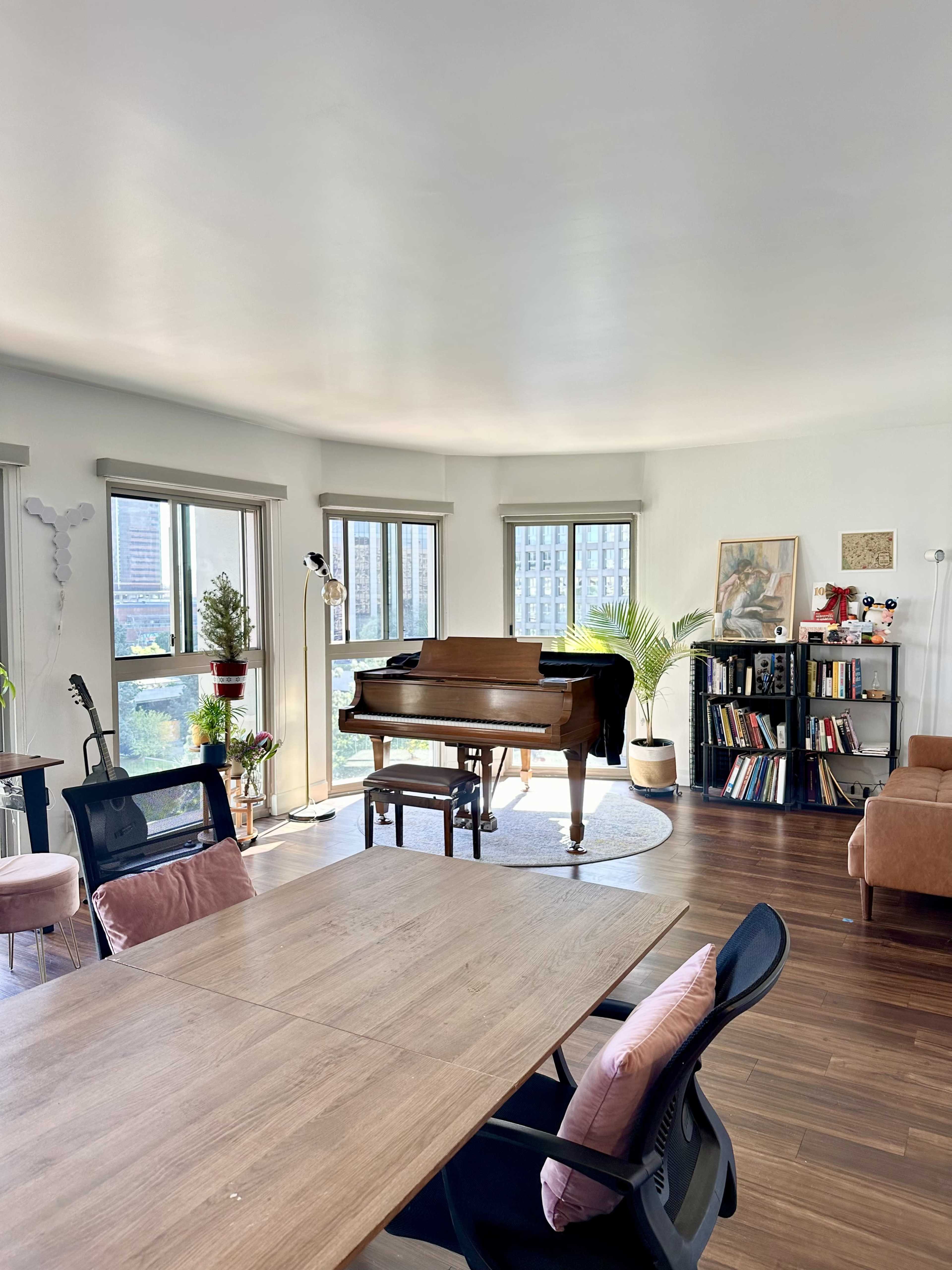 The image shows a bright, spacious living room featuring a grand piano in the center, surrounded by large windows and various pieces of furniture, including chairs, a bookshelf, and a guitar.
