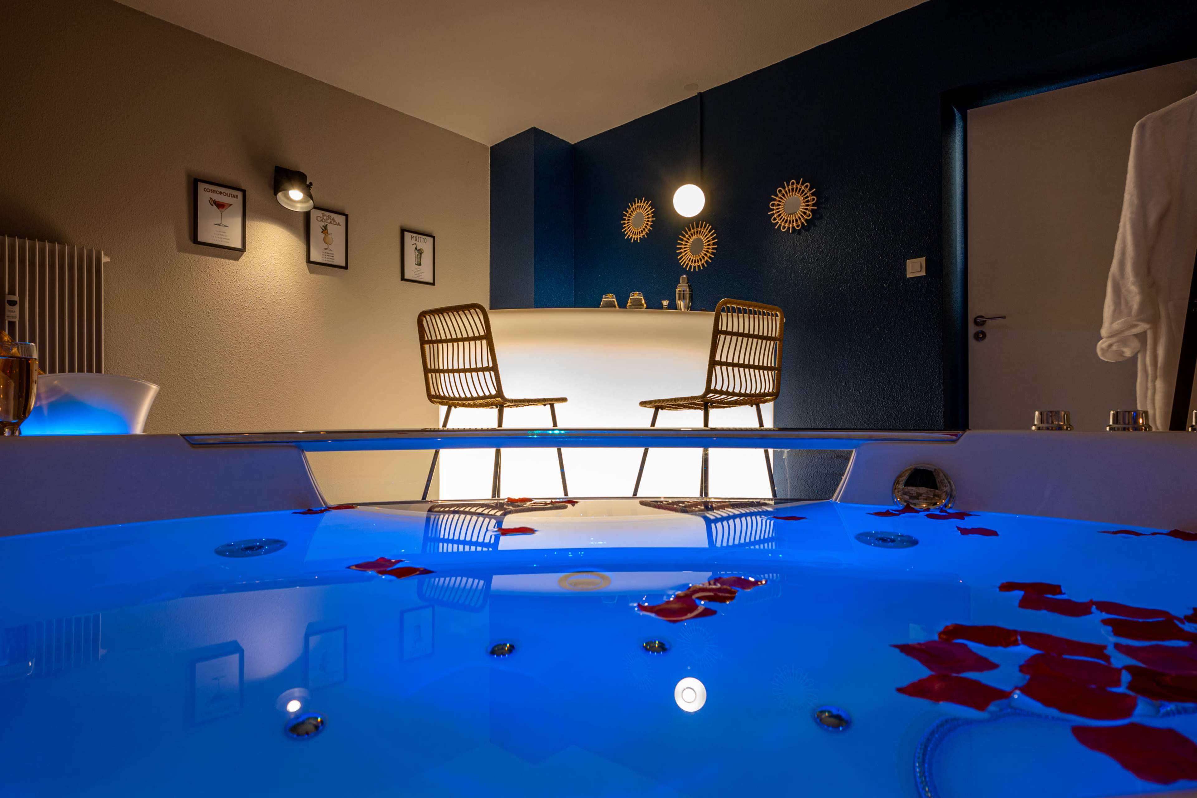 A modern bathroom features a jacuzzi with blue lighting and rose petals, while a minimalist bar with two chairs is in the background.