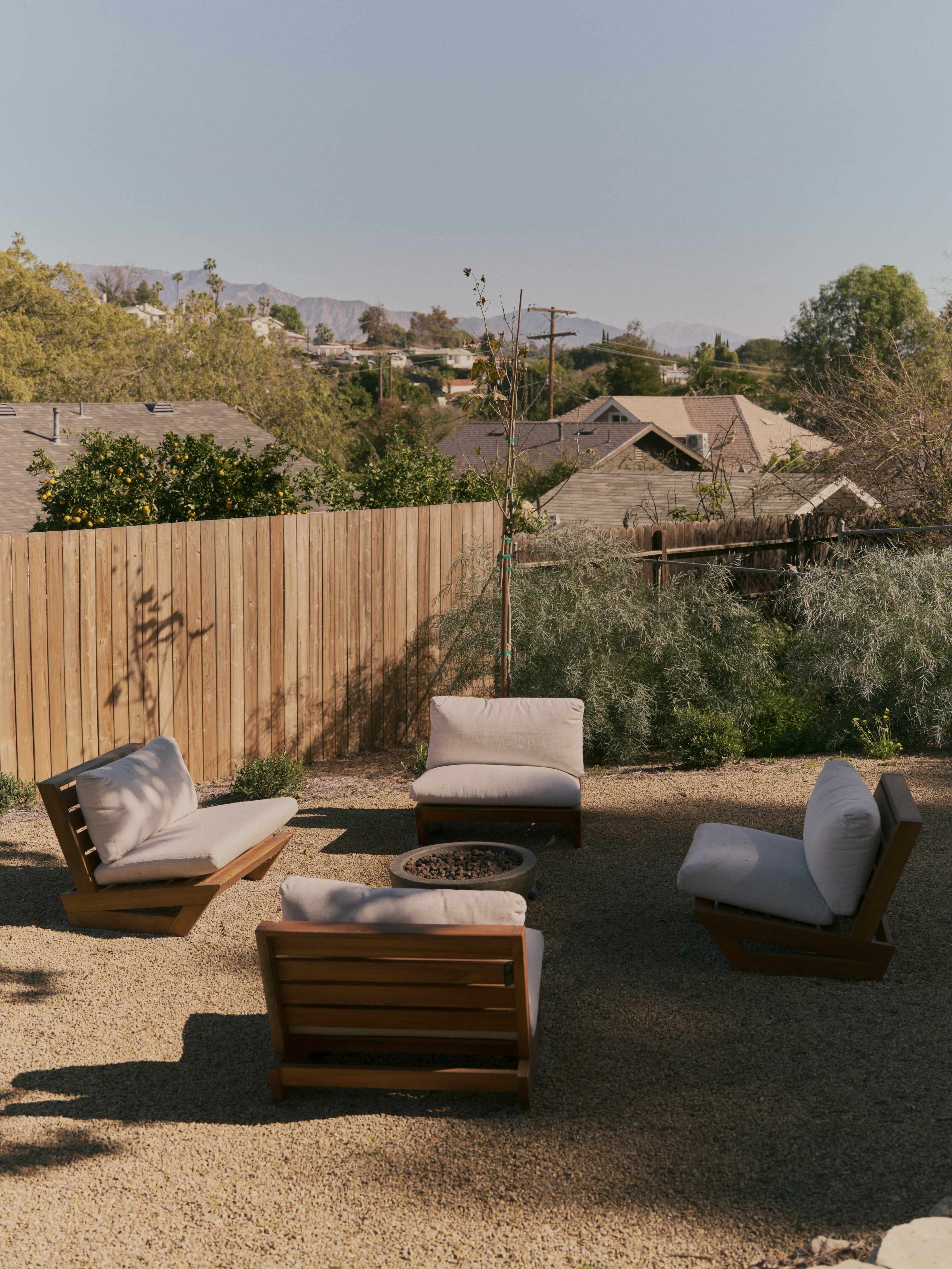 A circular seating arrangement with modern wooden chairs encircles a small fire pit in a gravel patio, surrounded by a wooden fence and trees in the background.