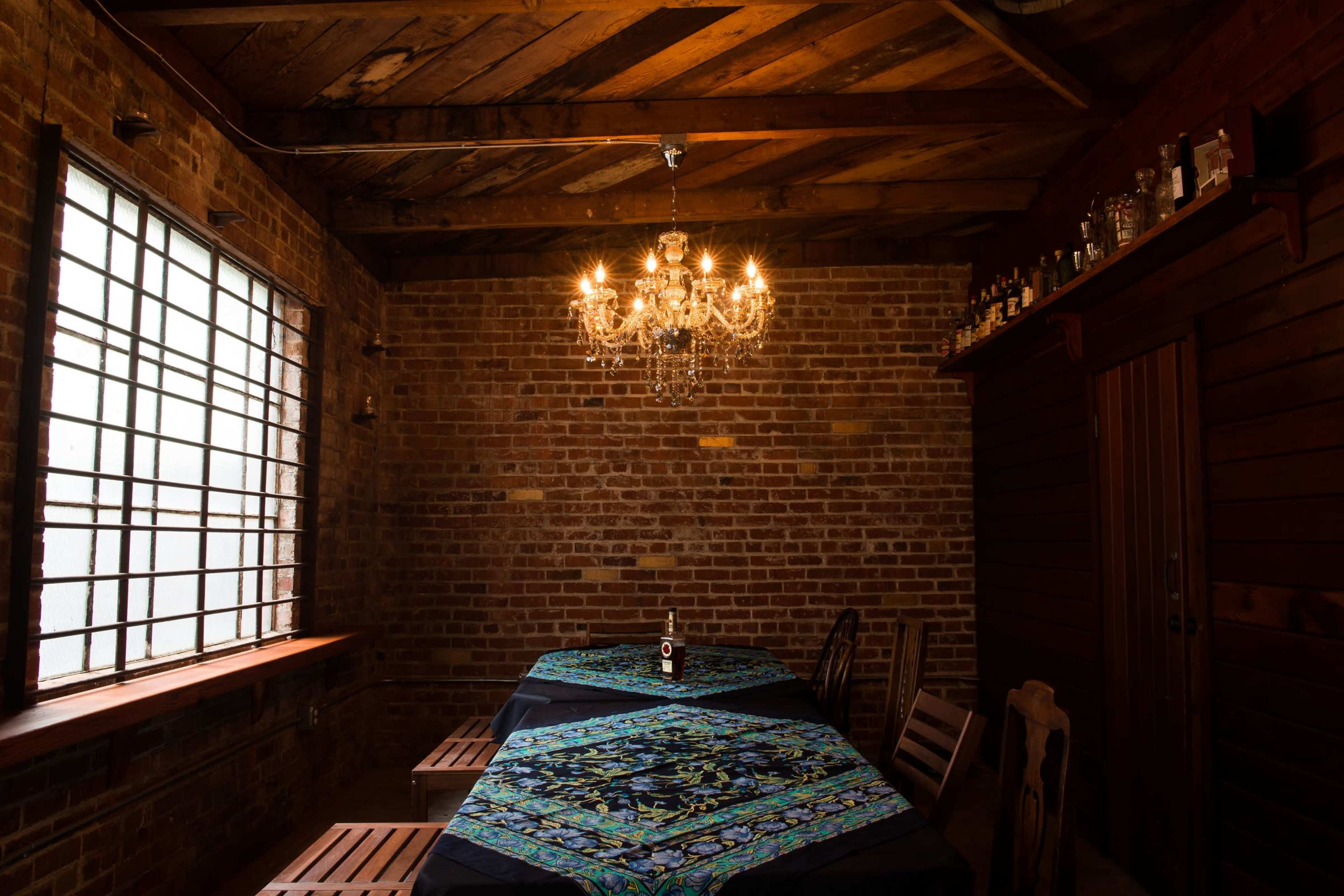 A wooden table is set beneath a chandelier in a brick-walled room with a window and shelves of bottles.