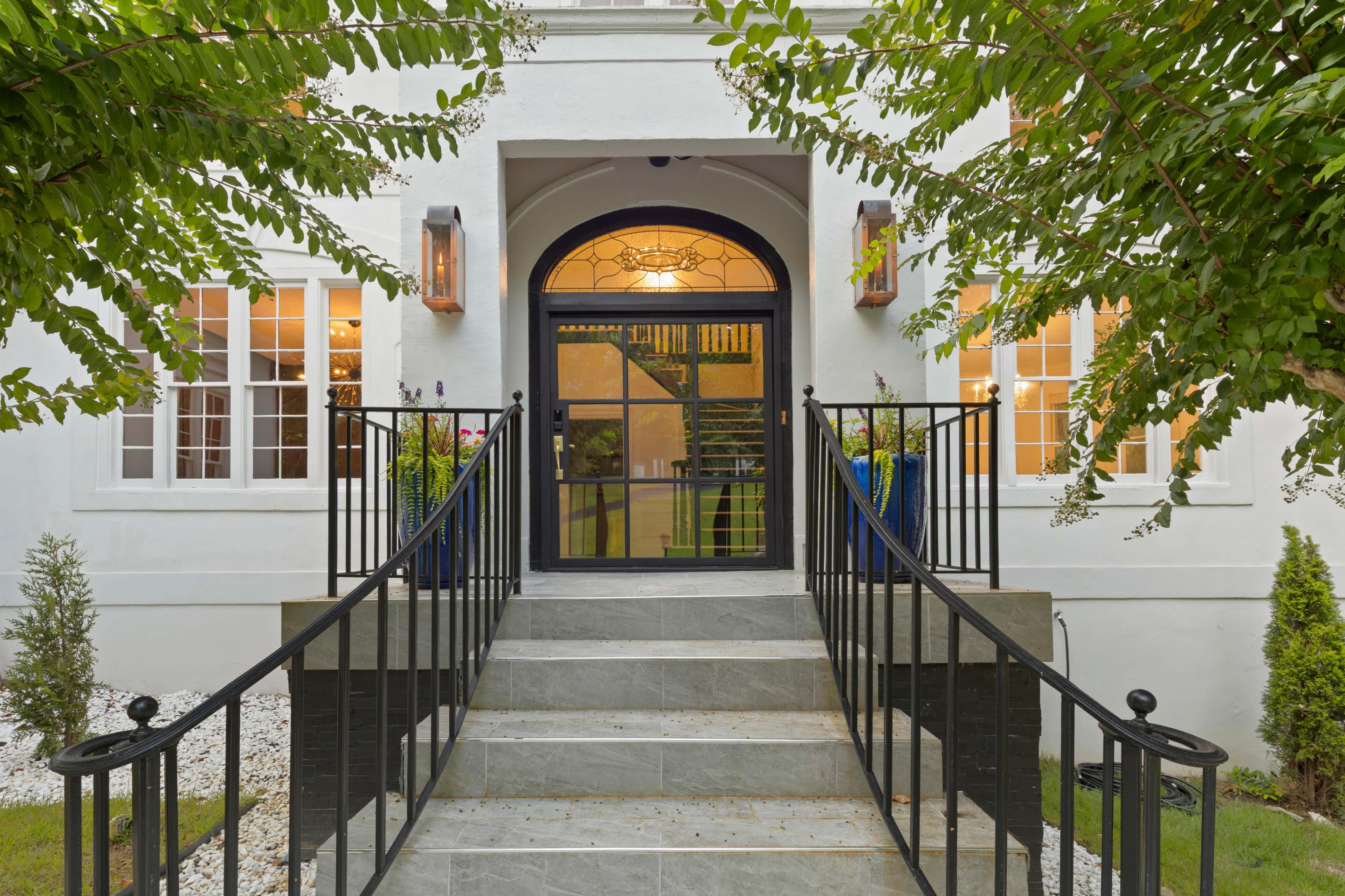 The image shows a modern house entrance with a black wrought iron railing and stairs leading to a large front door surrounded by greenery.