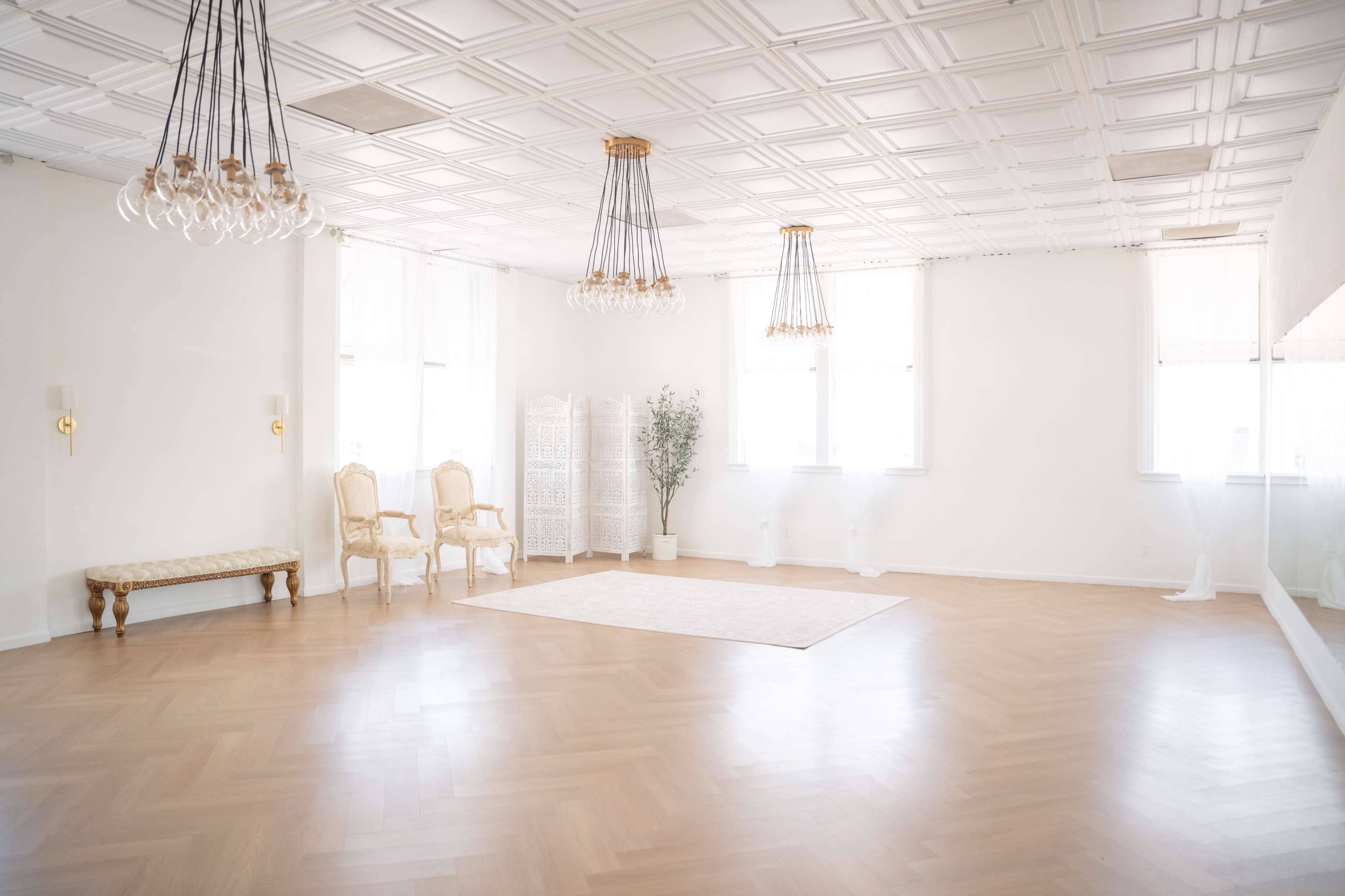 A bright, empty room with hardwood flooring, featuring ornate chandeliers and two elegant chairs near a decorative screen.