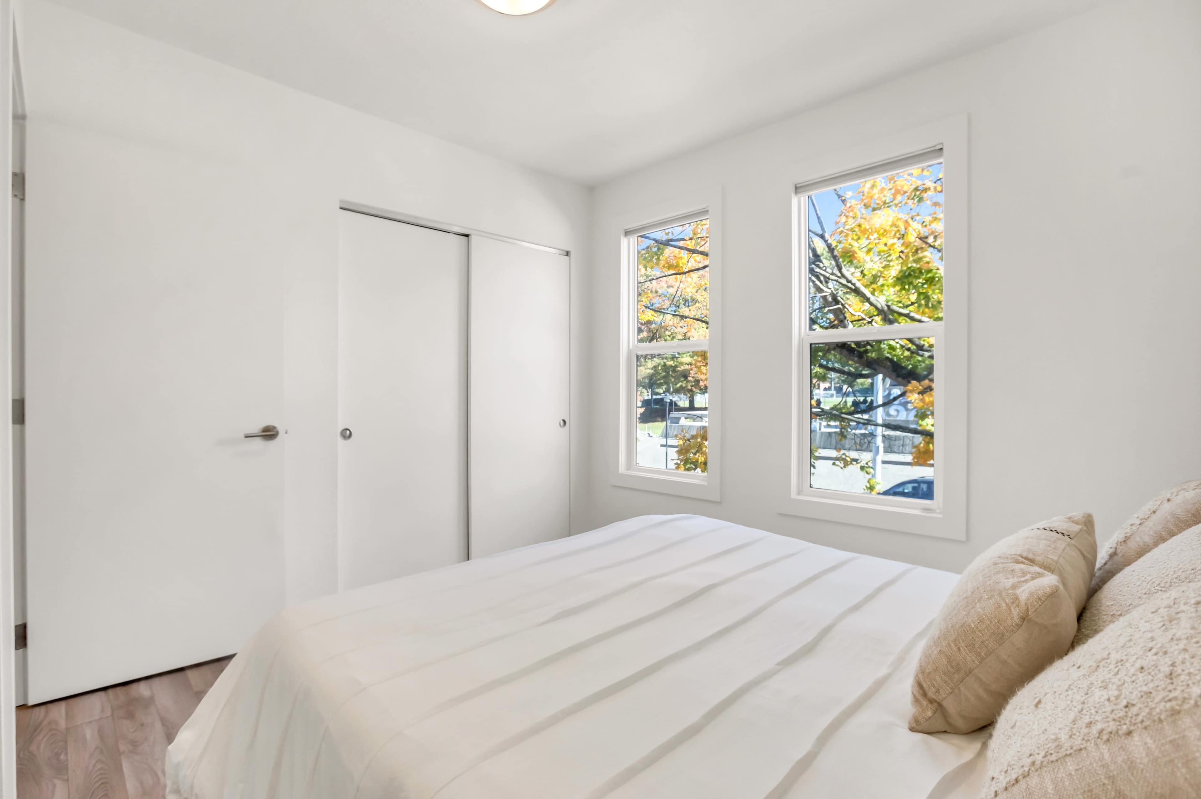 The image shows a bright, minimalist bedroom with a white bedspread, two windows, and a closet door.