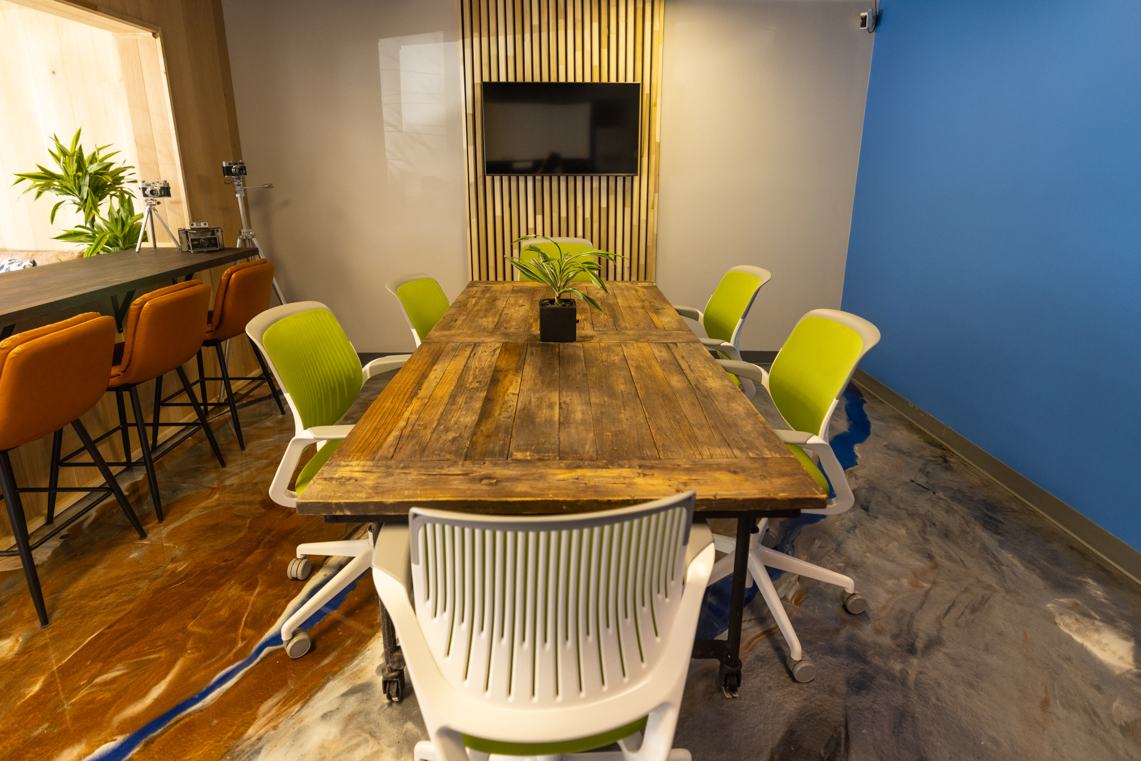 The image shows a modern conference room with a large wooden table surrounded by six chairs, a television mounted on the wall, and plants in the room.