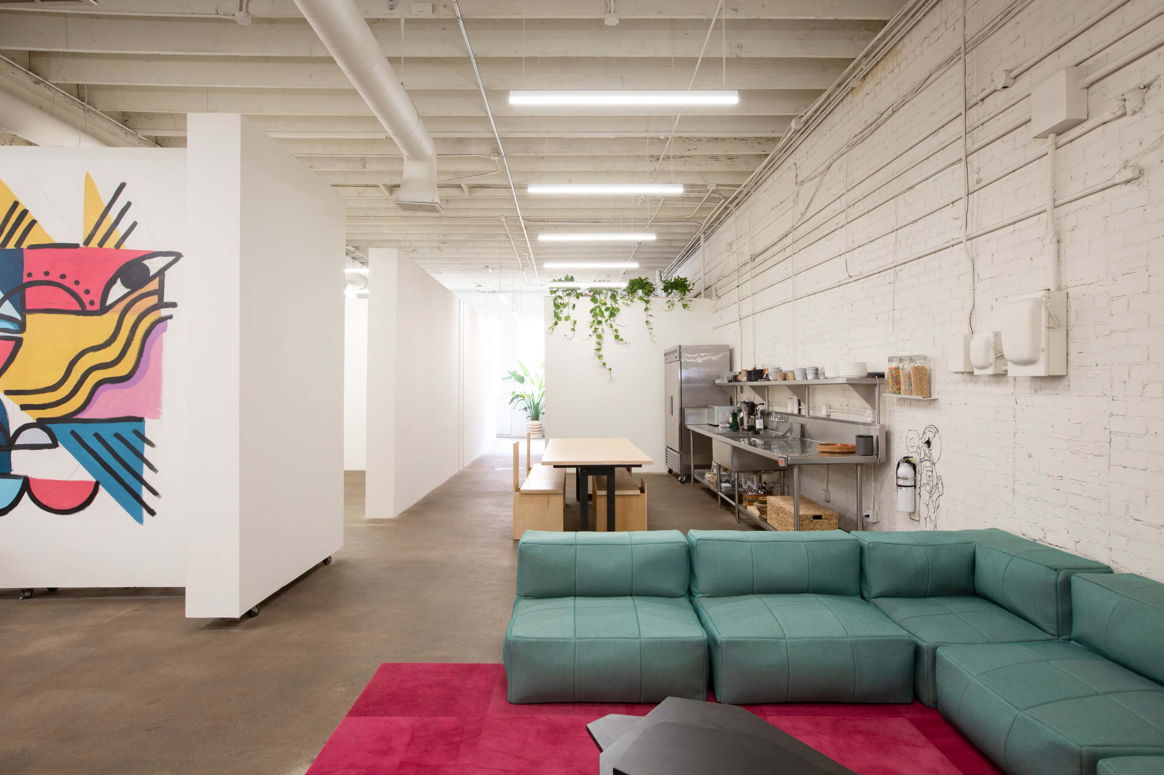 A modern, open interior space featuring a green sectional sofa, a pink rug, a long wooden table, and white walls adorned with a colorful mural.