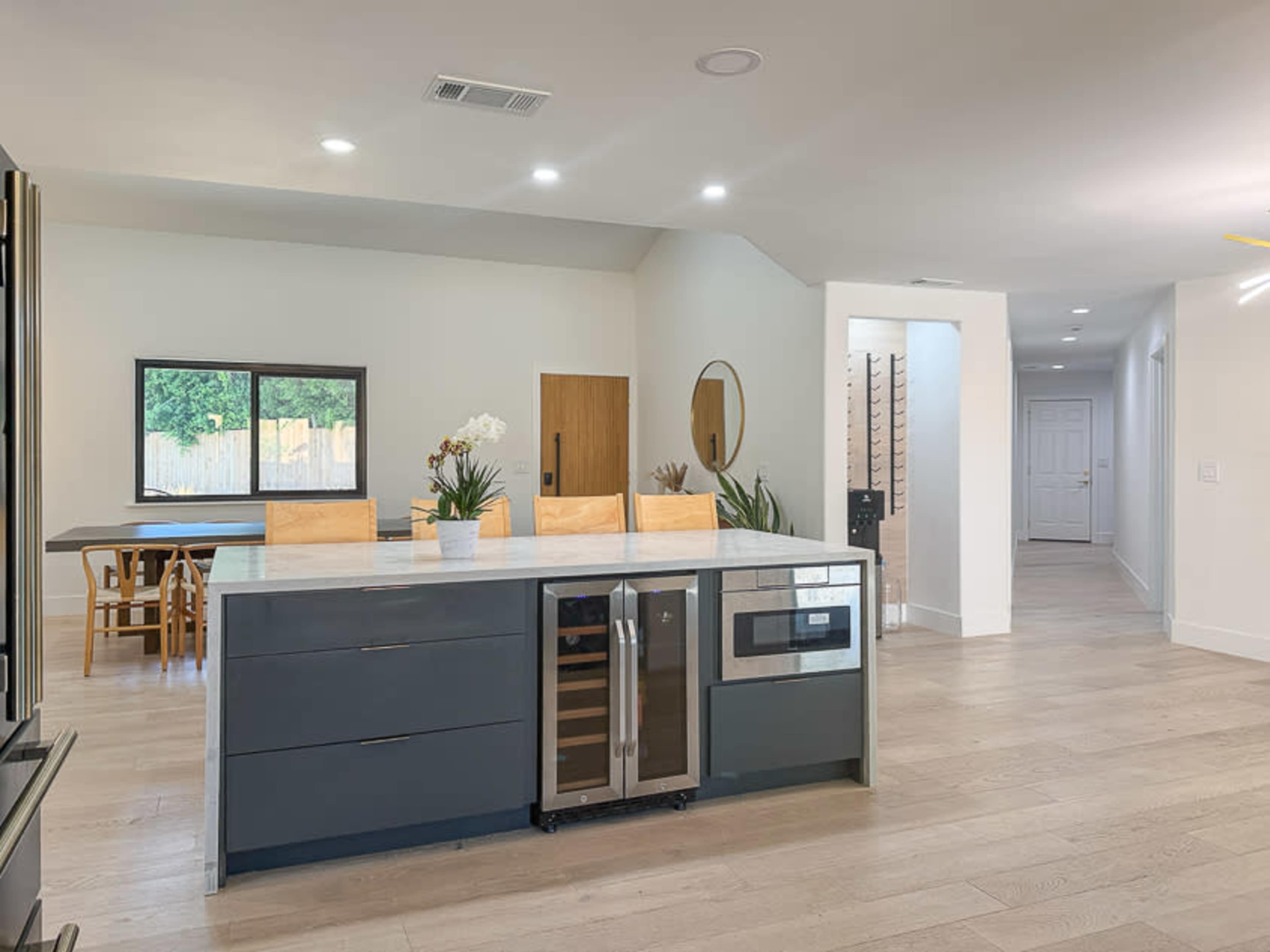 The image shows a modern kitchen with a central island featuring a built-in microwave and wine cooler, adjacent to a dining area with a wooden table and chairs in a well-lit, open layout.