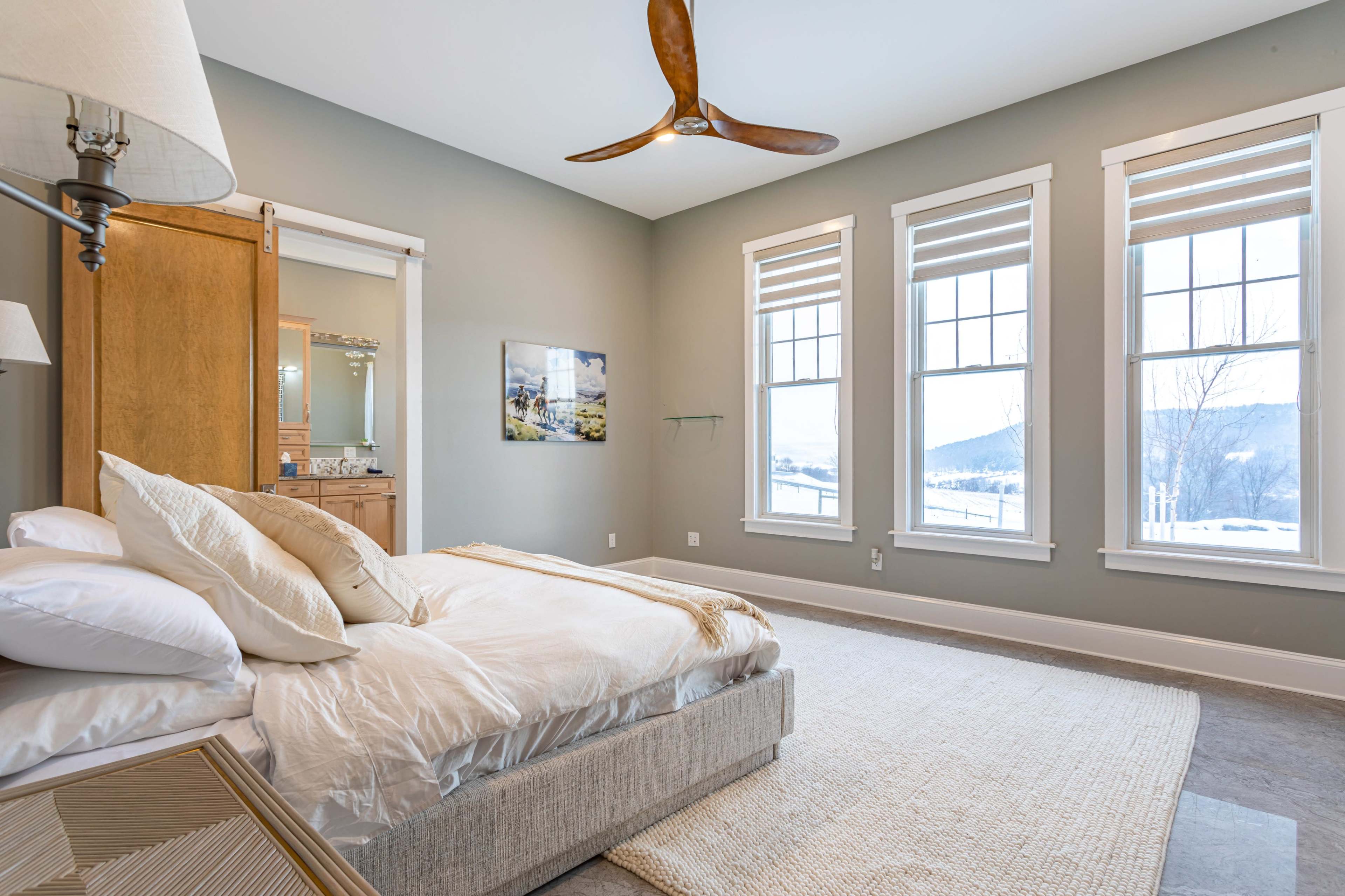 A modern bedroom features a bed with relaxed bedding, large windows with white trim, and a ceiling fan.