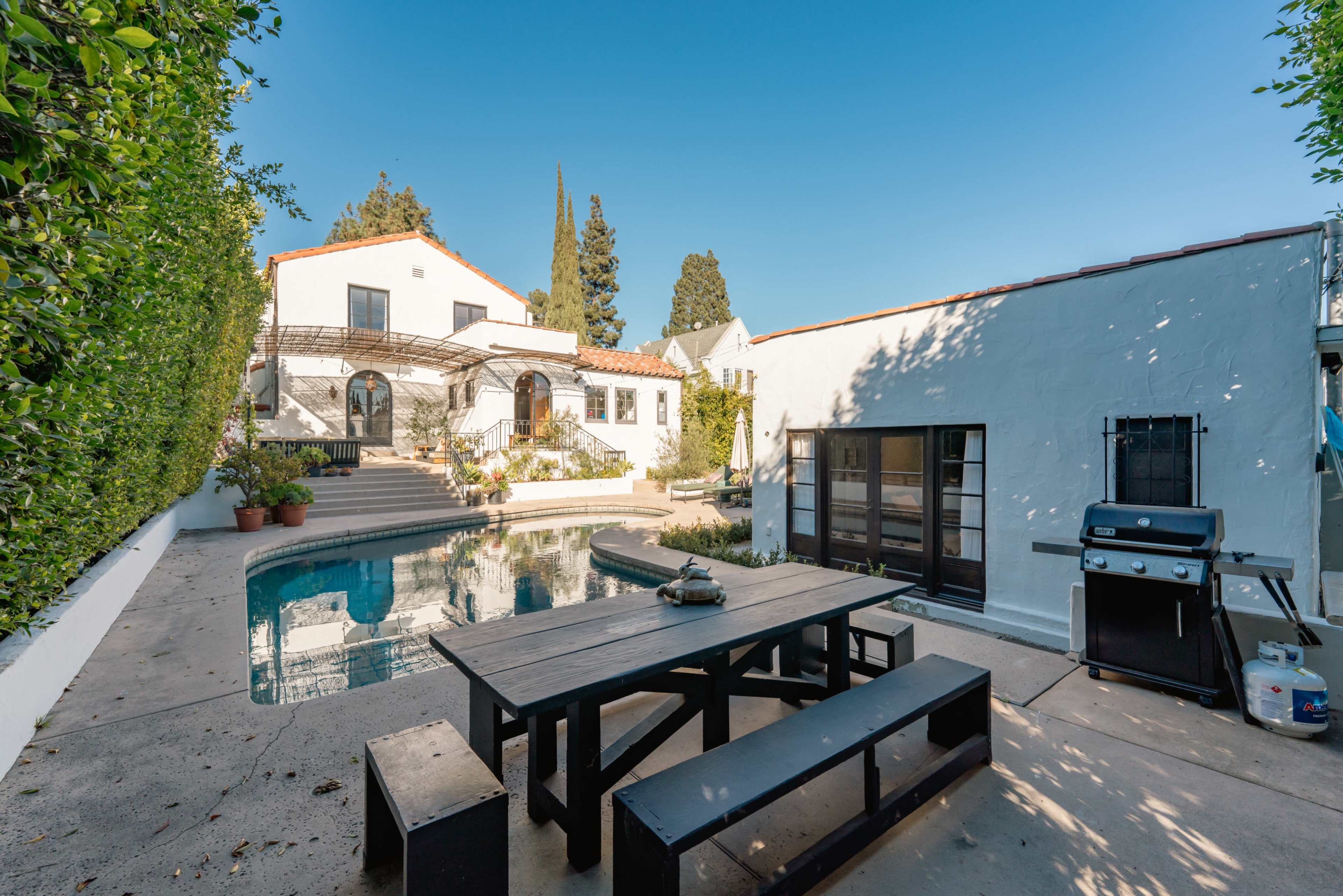 The image shows a courtyard with a swimming pool, surrounded by white buildings and a black outdoor dining table with benches.