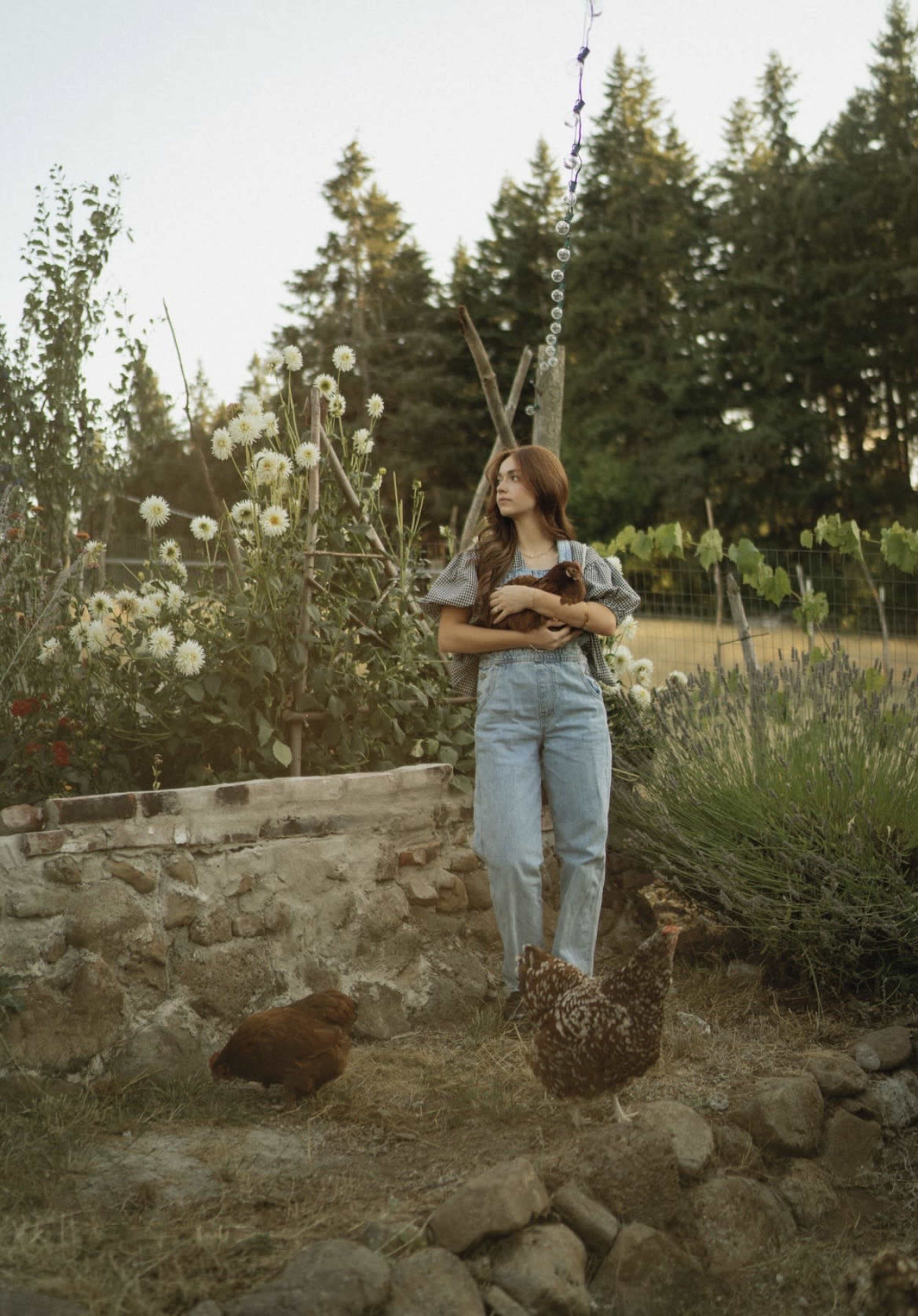 A woman stands in a garden with flowers and chickens, holding a small animal, surrounded by trees in the background.