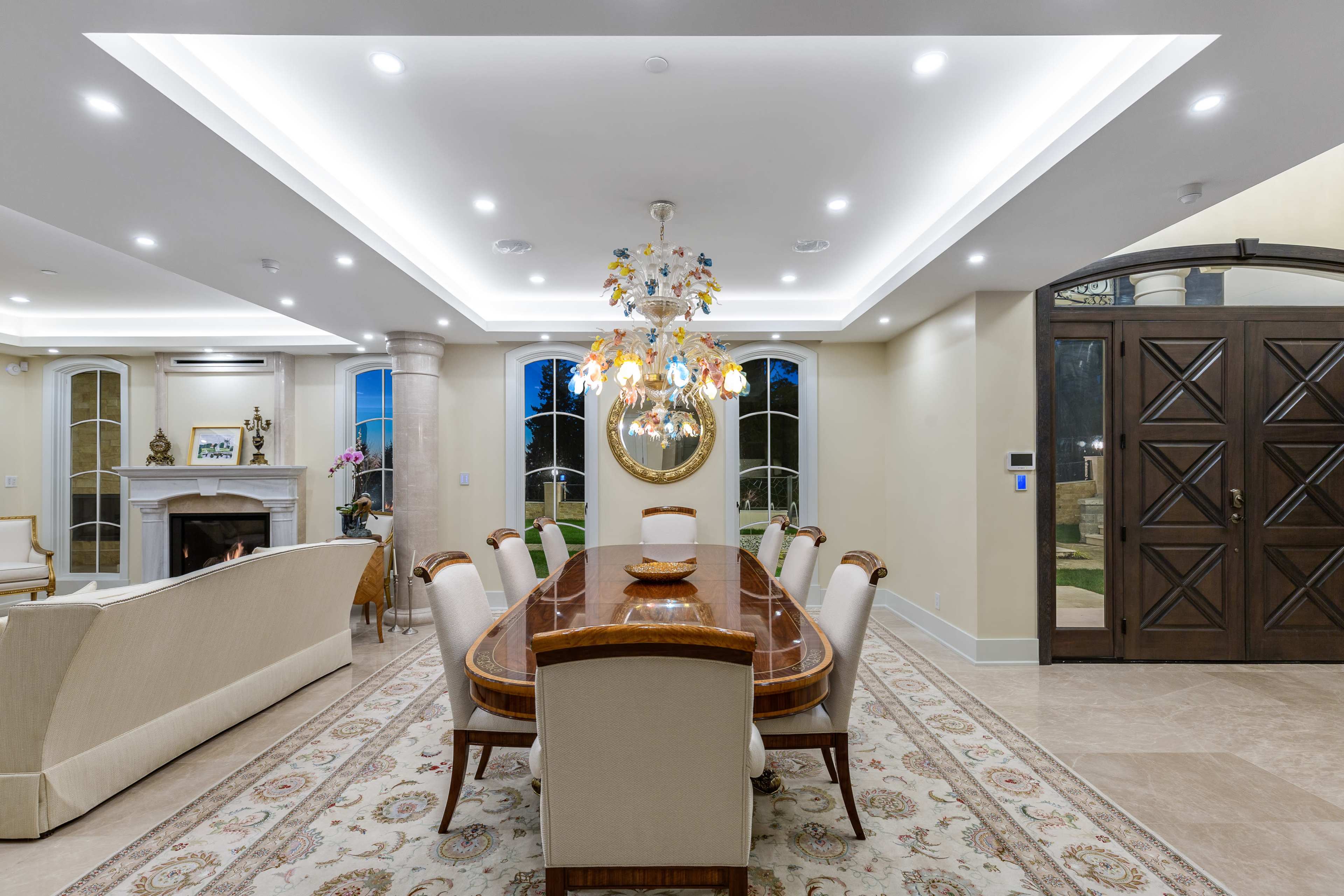 The image shows a spacious dining room featuring a long wooden table surrounded by upholstered chairs, with a decorative chandelier hanging above and a fireplace in the background.