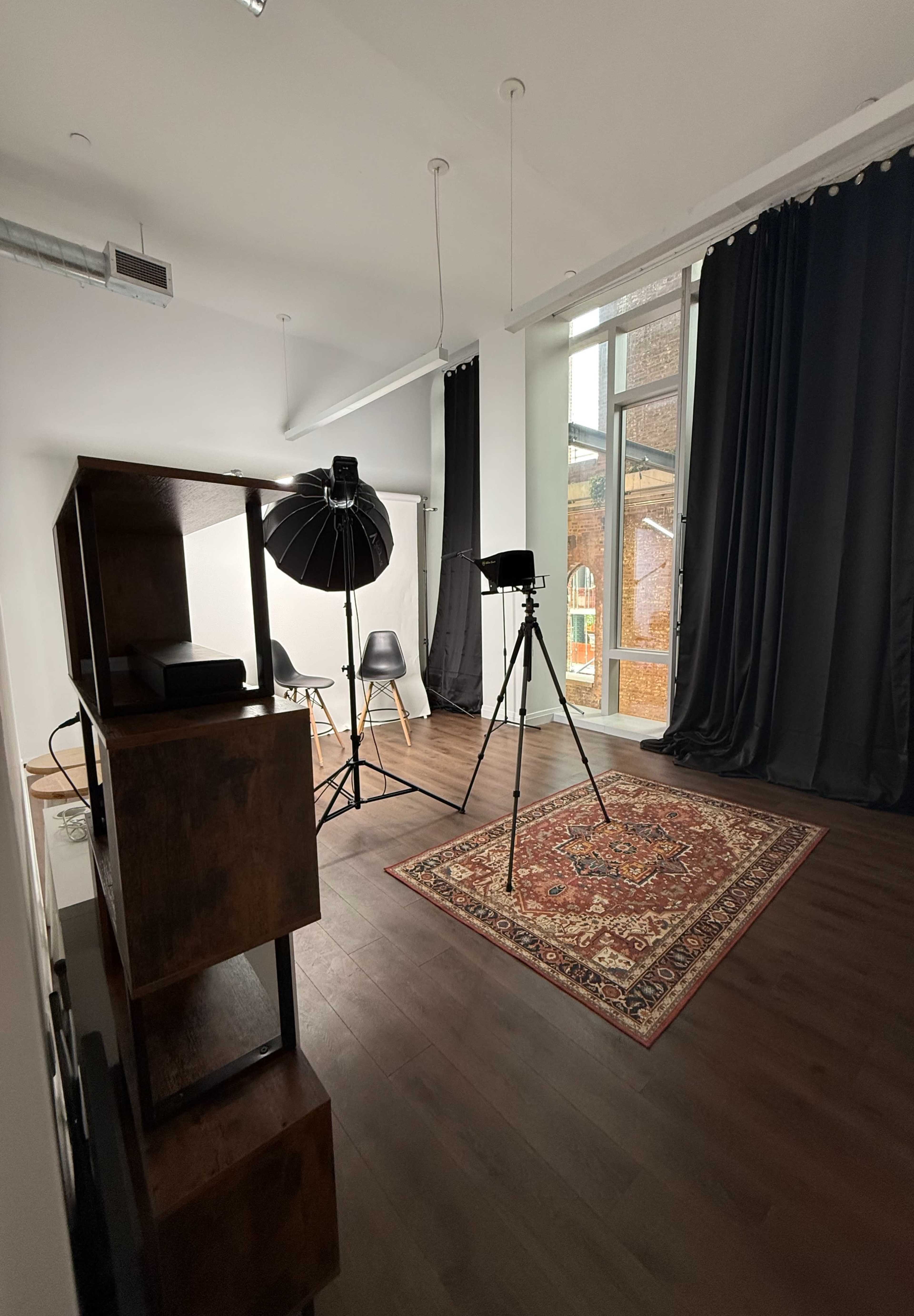 The image shows a photography studio featuring a black backdrop, a lighting setup, a tripod, and a wooden bookshelf in a well-lit room with large windows.
