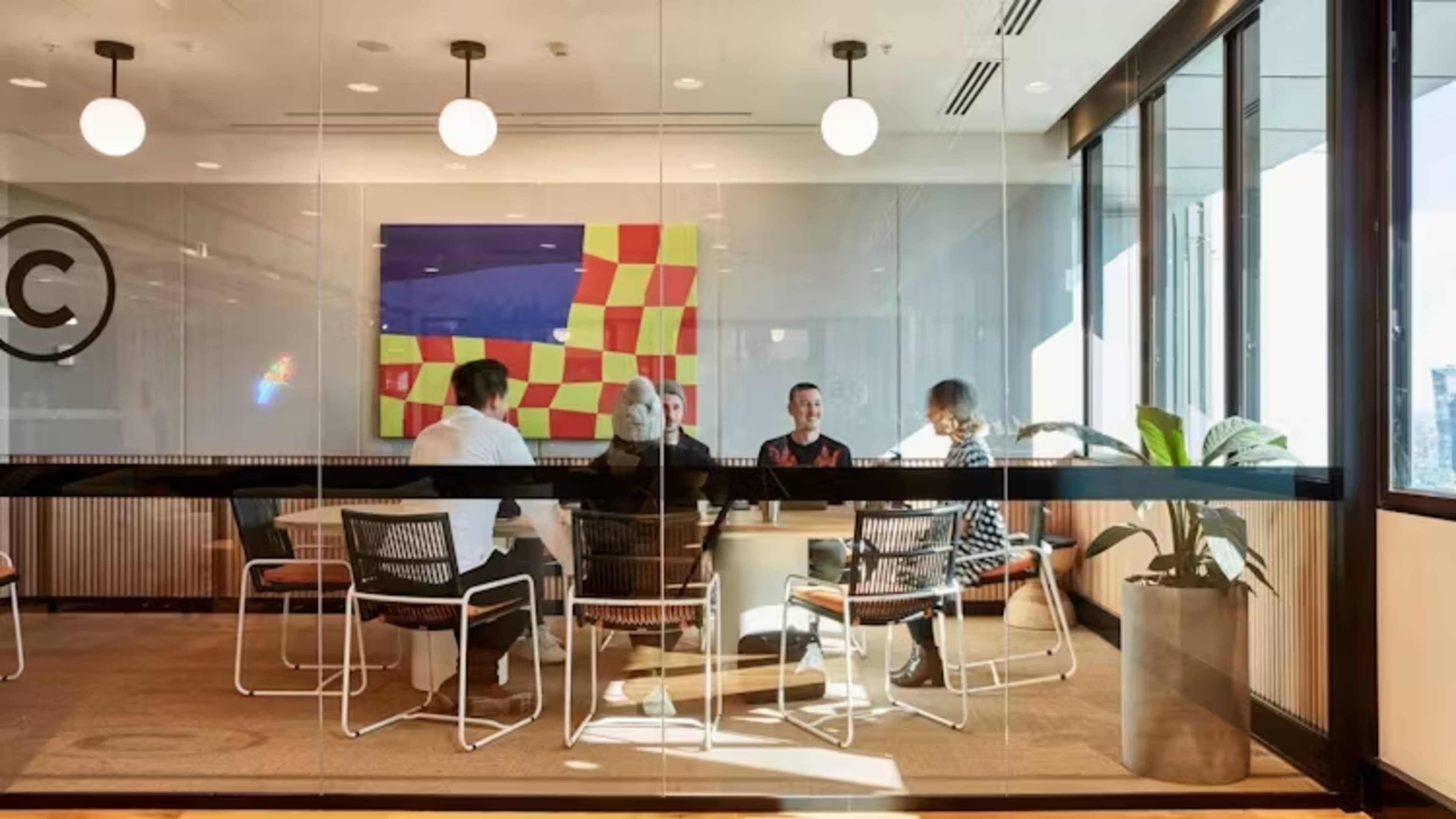 A group of four people is seated around a table in a glass-walled conference room, with colorful artwork visible on the wall.